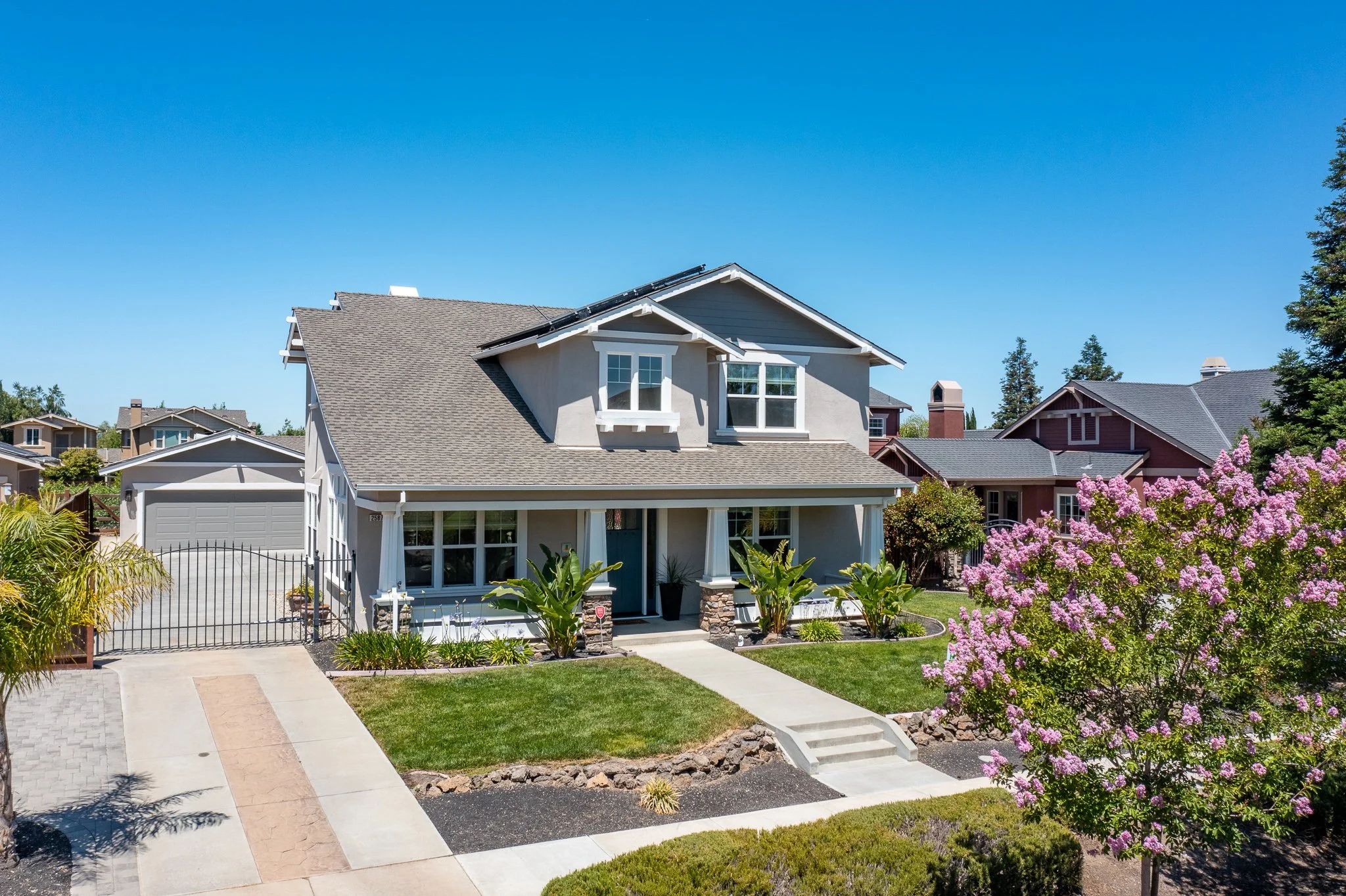 Front view of a modern two-story house with a manicured lawn, pink flowering tree, and neighboring houses against a blue sky.