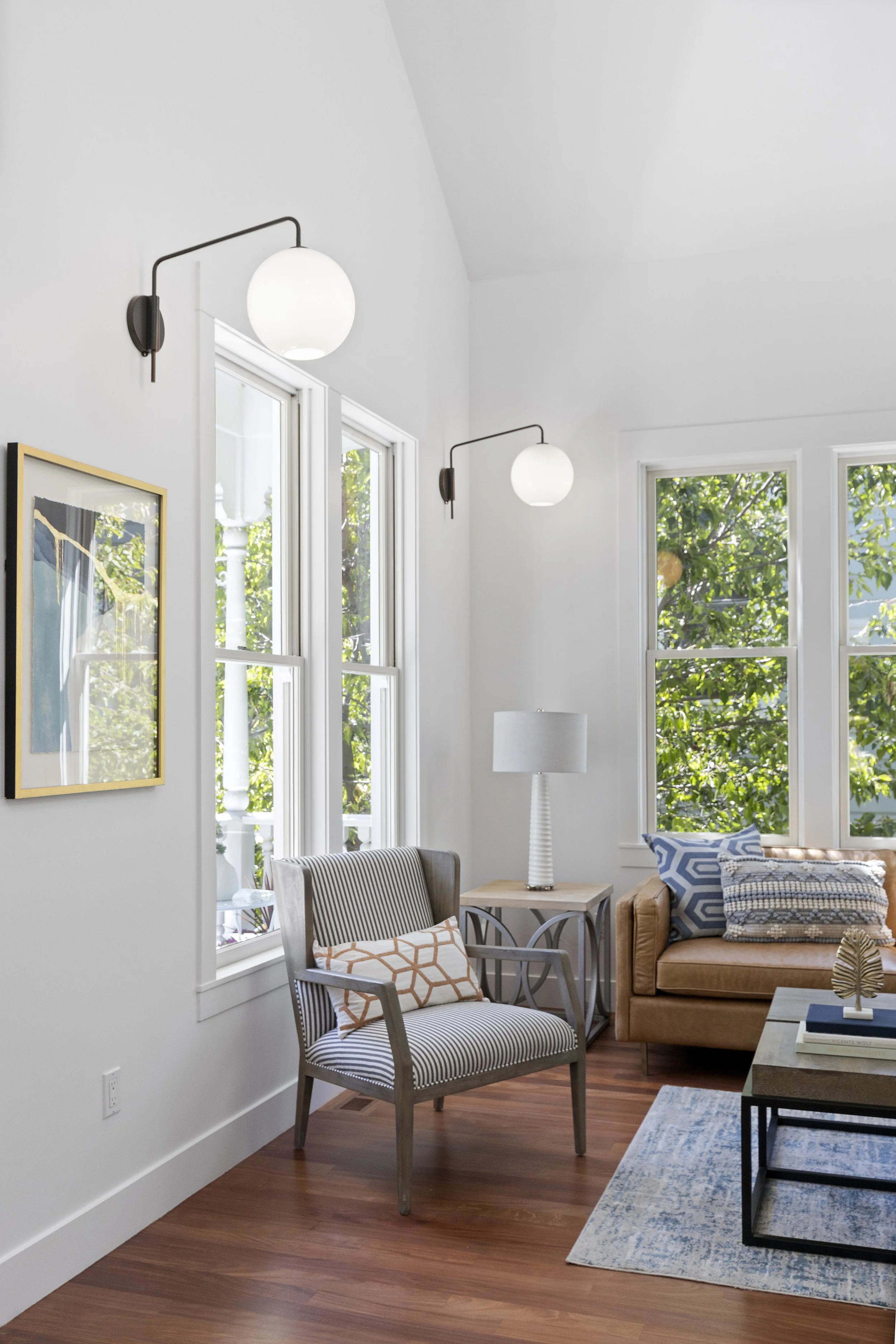 Living room with white walls, large windows, a brown leather sofa with patterned pillows, a striped armchair with an orange geometric pillow, a side table with a white lamp, some books, a blue patterned rug, and modern wall sconces with globe lights.