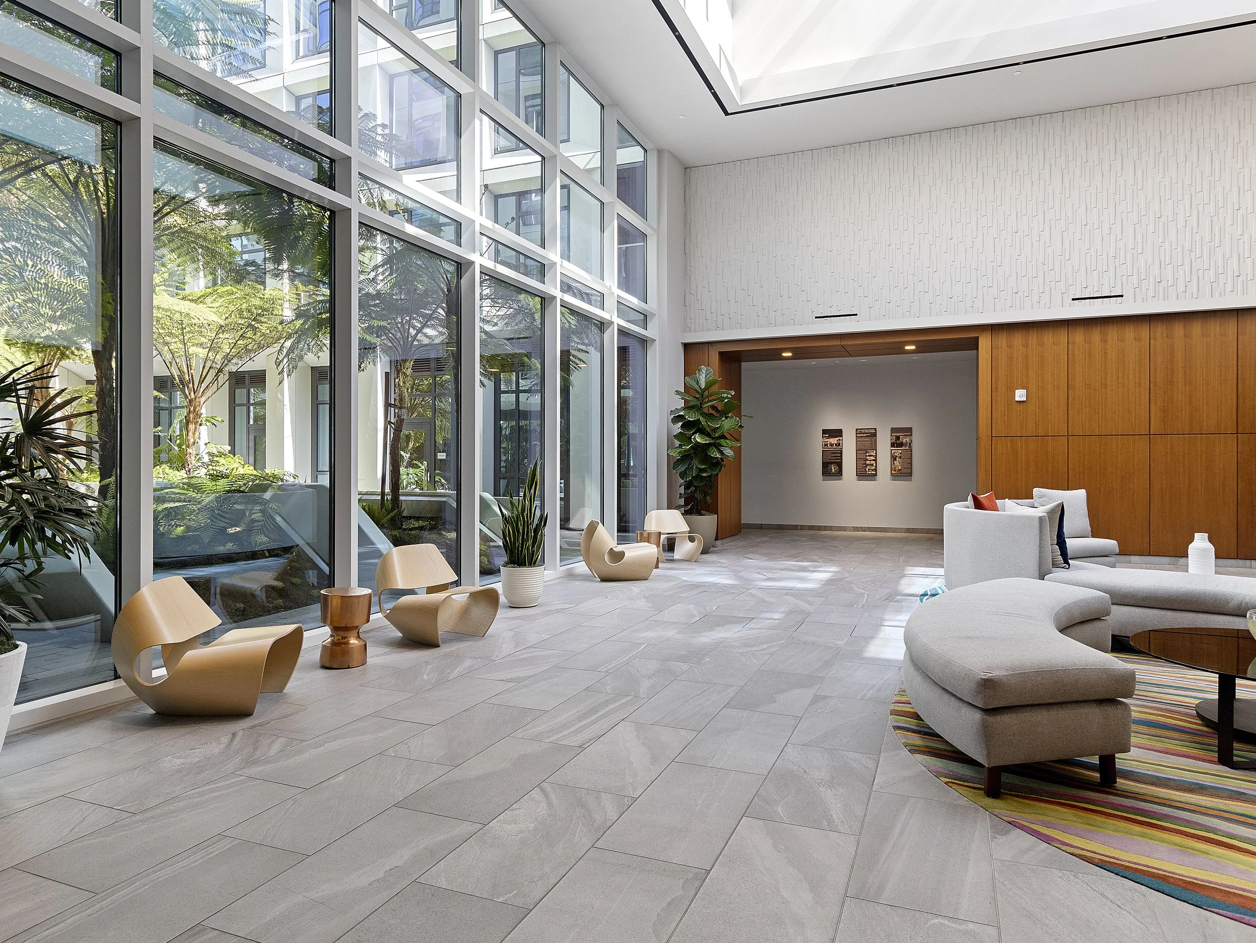 Modern indoor space with large floor-to-ceiling windows, sitting area with armchairs, sofas, wooden and ceramic furniture, indoor plants, and a colorful striped rug.