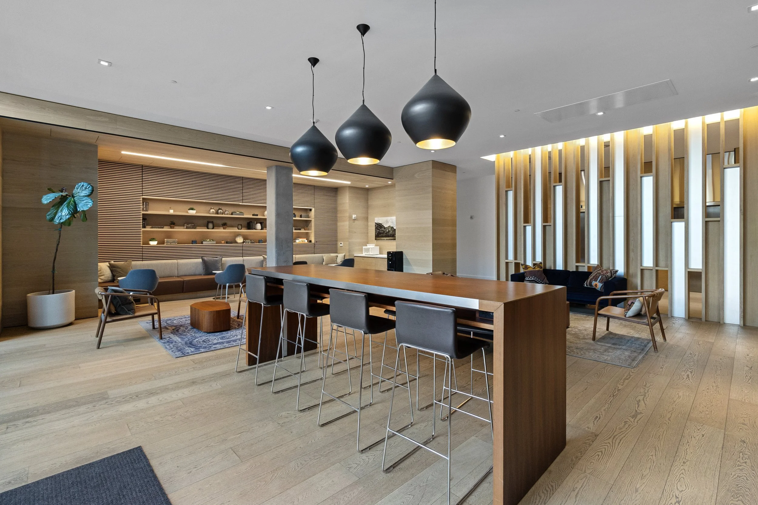 Interior of a modern lounge or community space with wooden flooring, black hanging pendant lights, a sitting area with chairs and couches, and a decorative wooden partition.