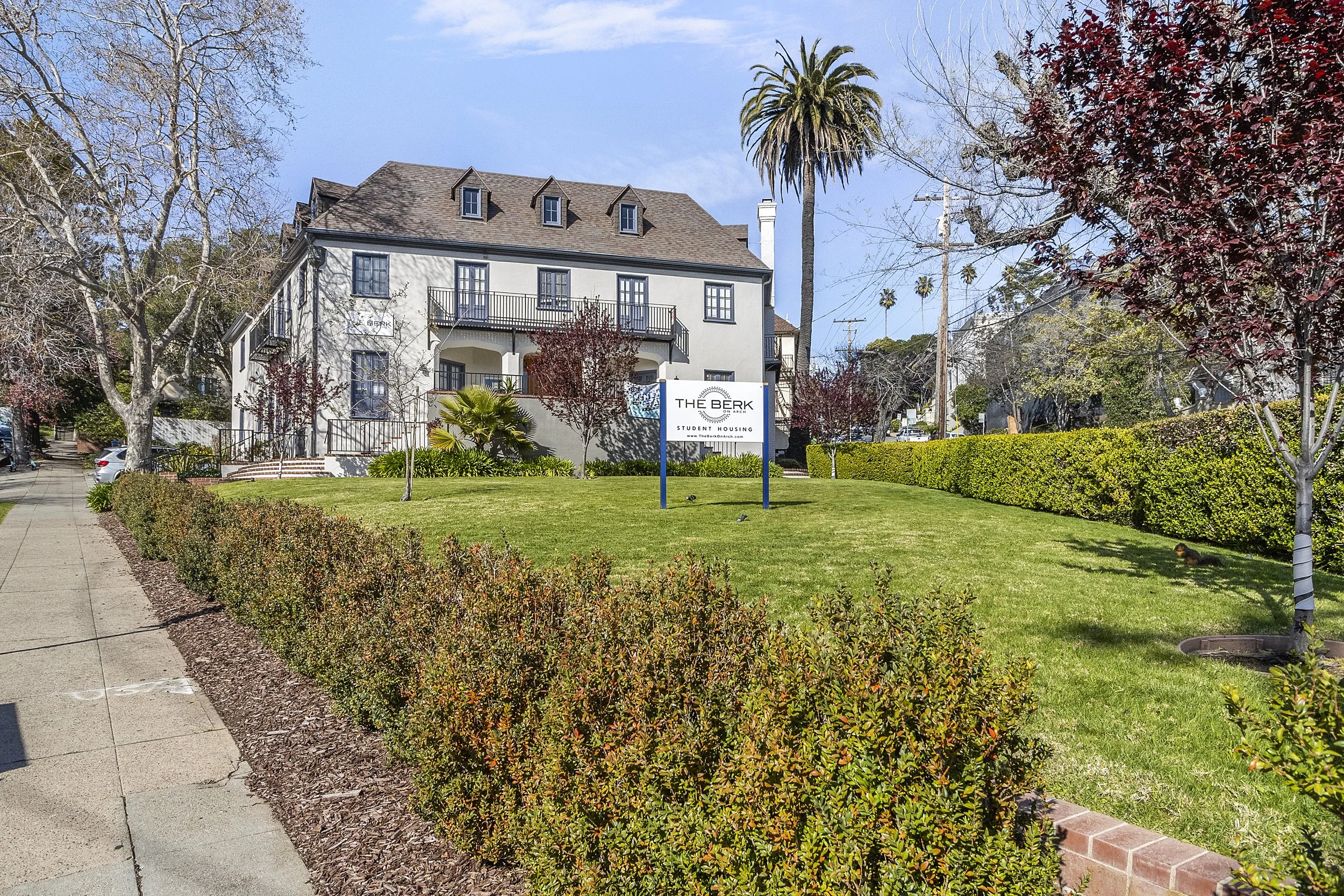 A multi-story white building with a brown gabled roof, labeled 'The Berk Student Housing,' is surrounded by trees and a well-maintained lawn with a sign in the front. The scene includes a sidewalk, some parked cars, and a partly cloudy sky.
