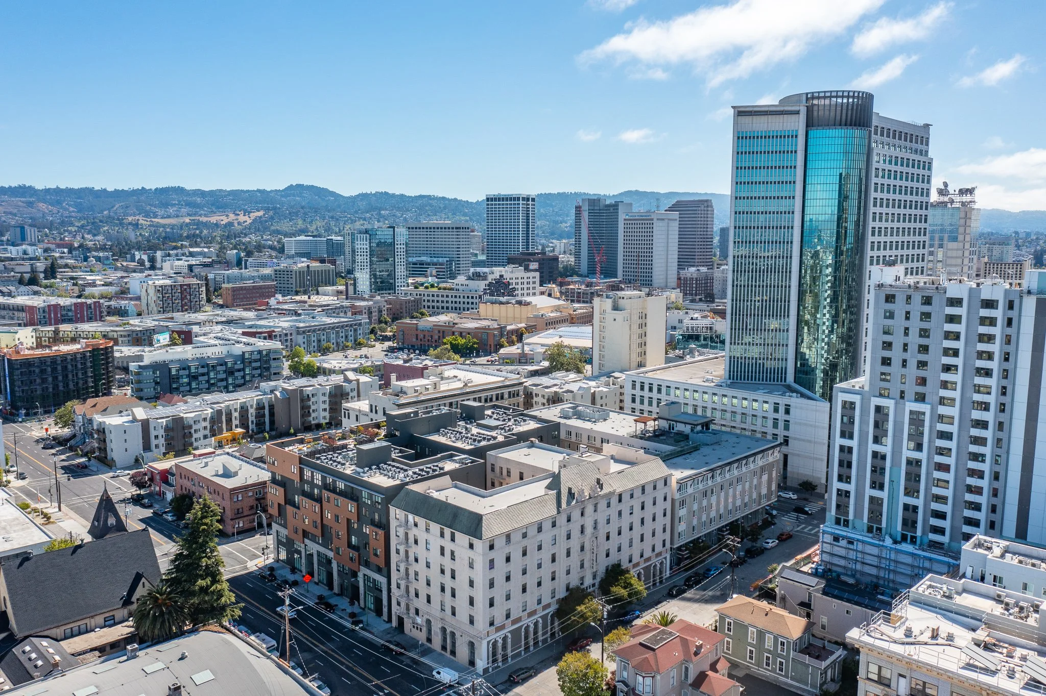 A high-angle view of a cityscape showing tall modern buildings, apartment complexes, and streets, with a mountain range in the background and a partly cloudy sky overhead.