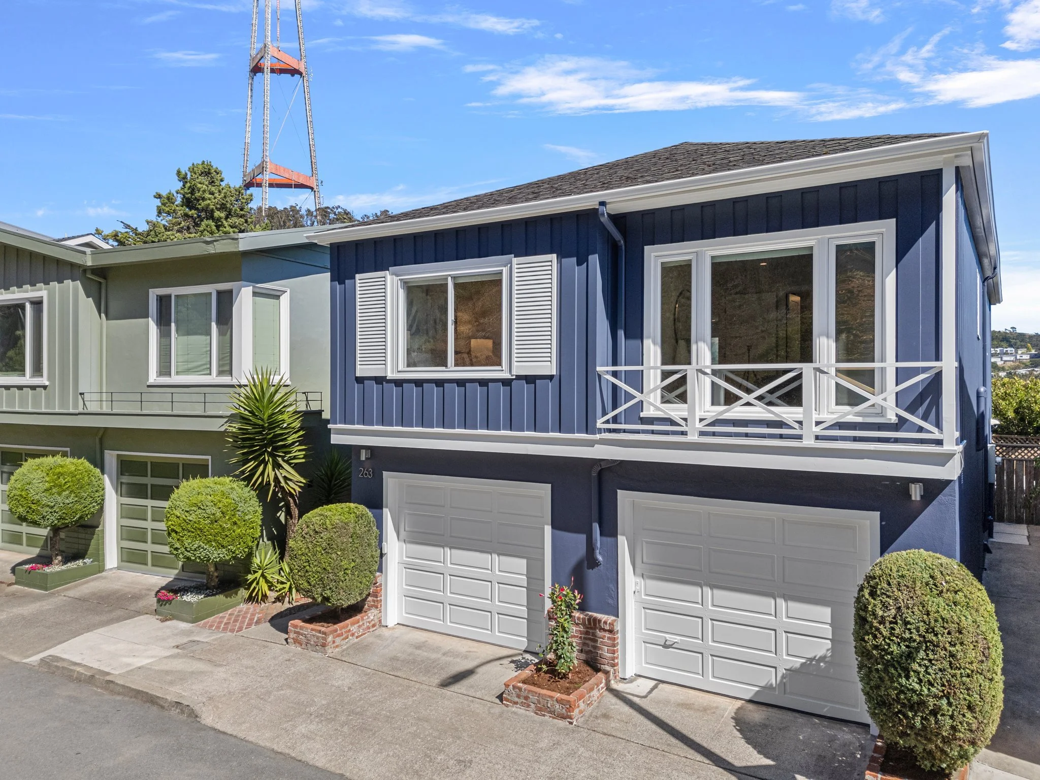 Two attached houses with the lower parts painted in light green and upper parts in dark blue, each with white garage doors and windows. There are small landscaped areas with bushes and plants in front of each house, and a concrete driveway and sidewa