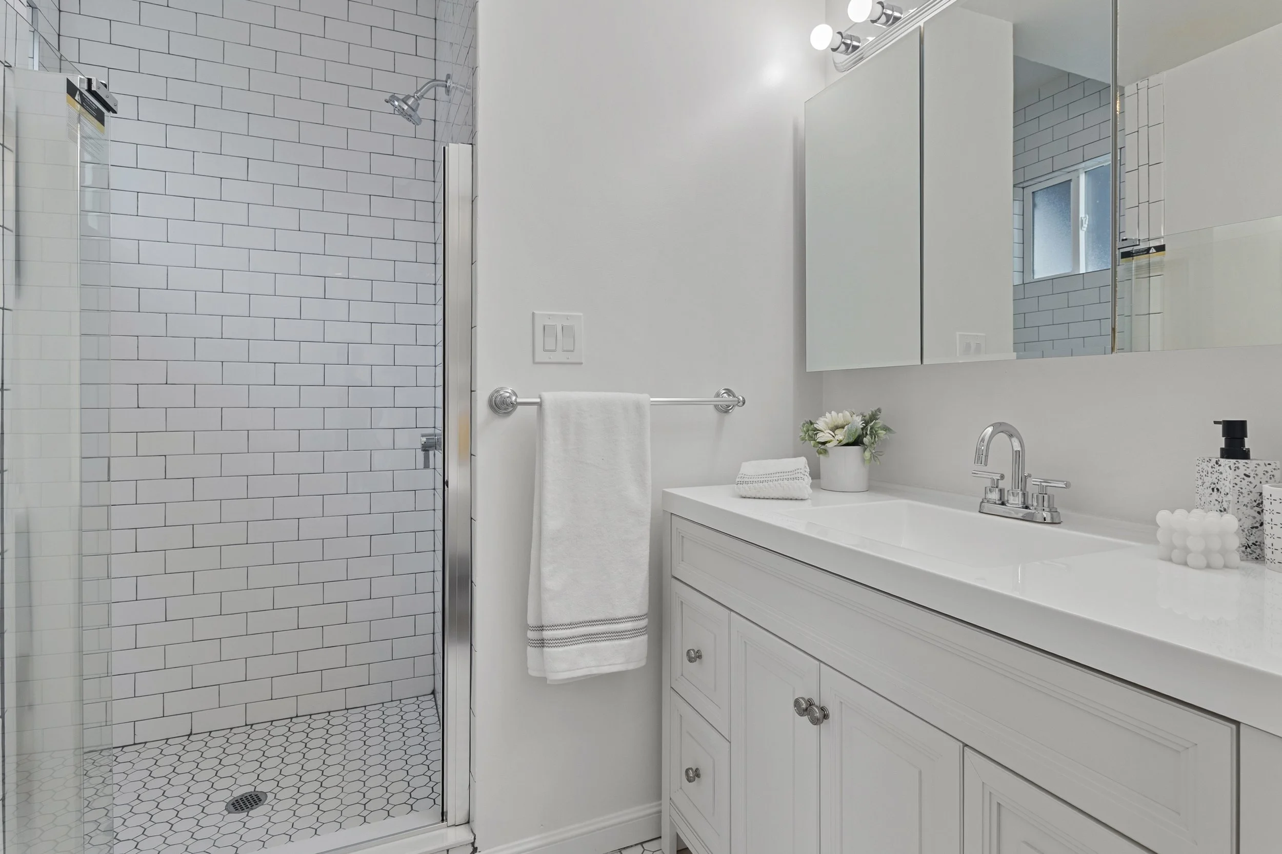 Bathroom with white subway tile shower, white vanity with silver handles, large mirror, small window, flower arrangement, soap dispenser, towel hanging on towel bar.
