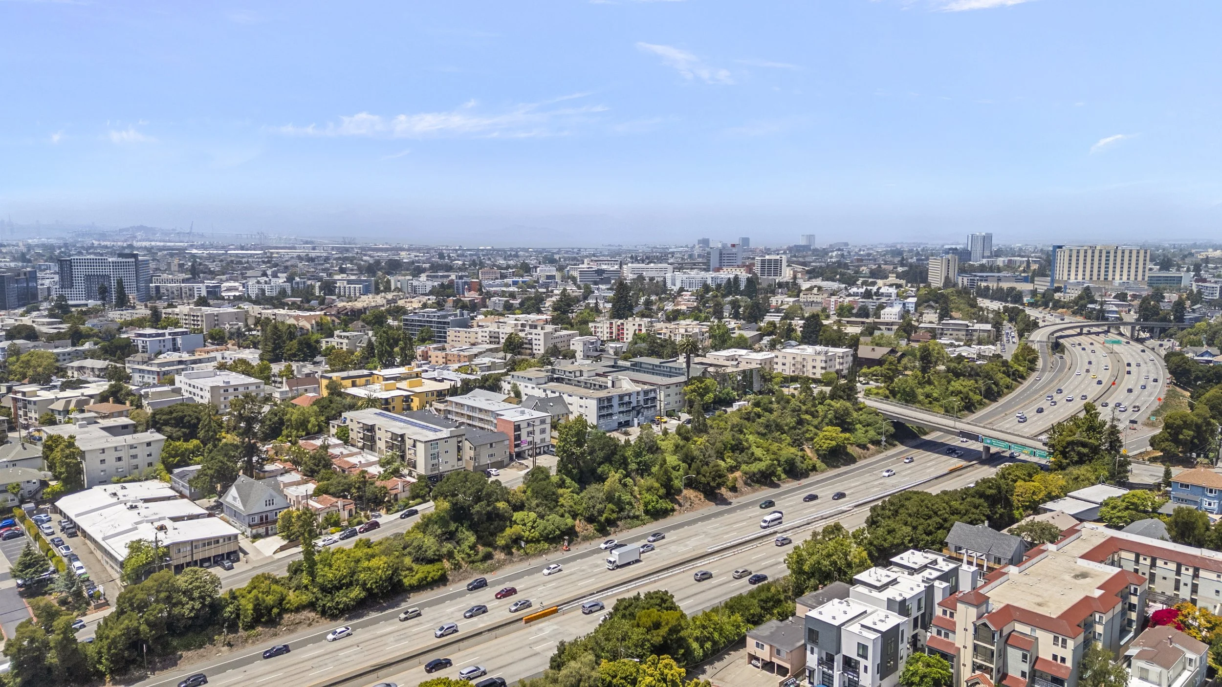 Aerial view of a cityscape with residential and commercial buildings, highways, and green trees under a blue sky with some clouds.