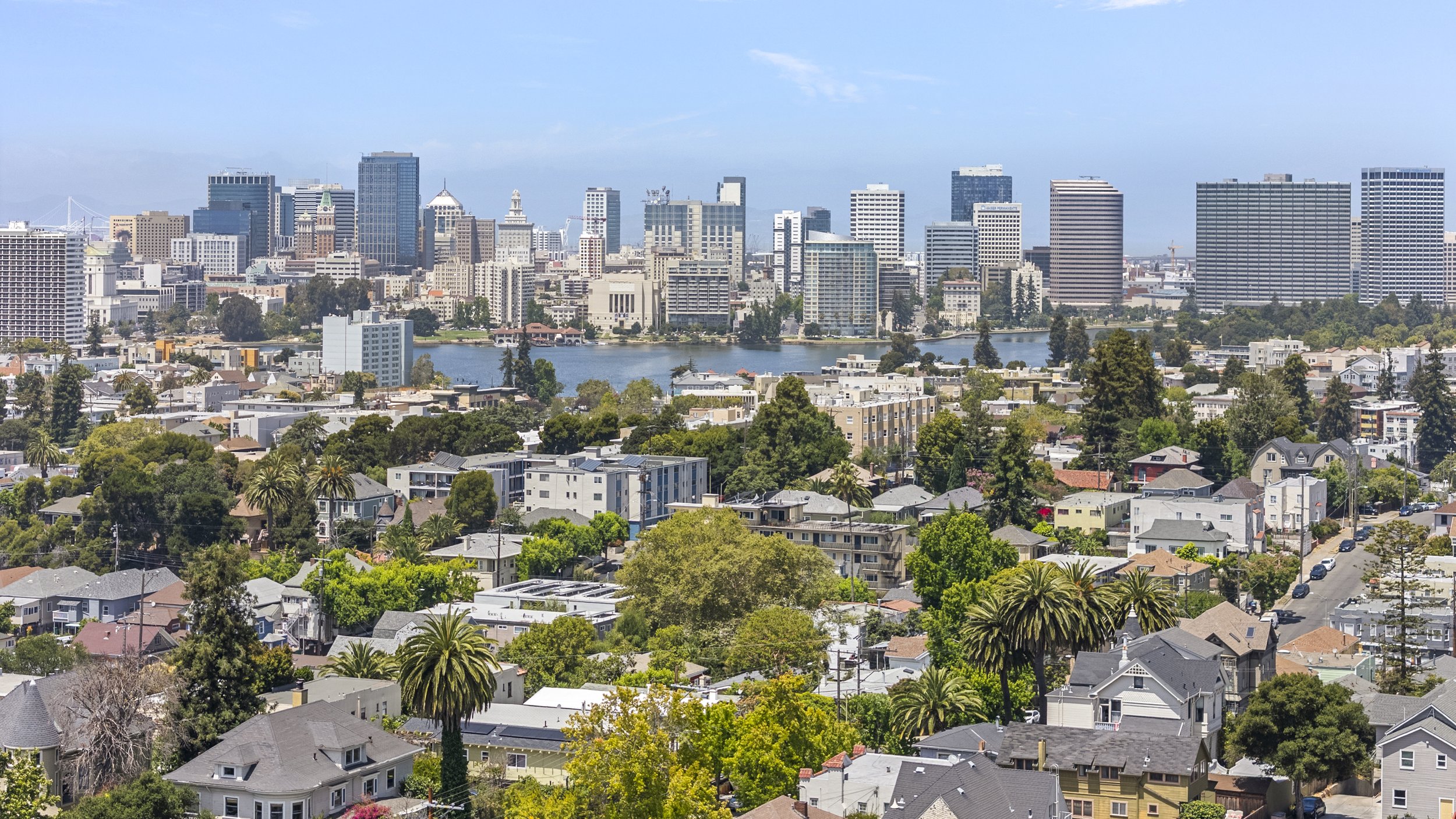 A city skyline with tall modern skyscrapers and a pond in the foreground, surrounded by residential neighborhoods with many trees and houses, under a clear blue sky.