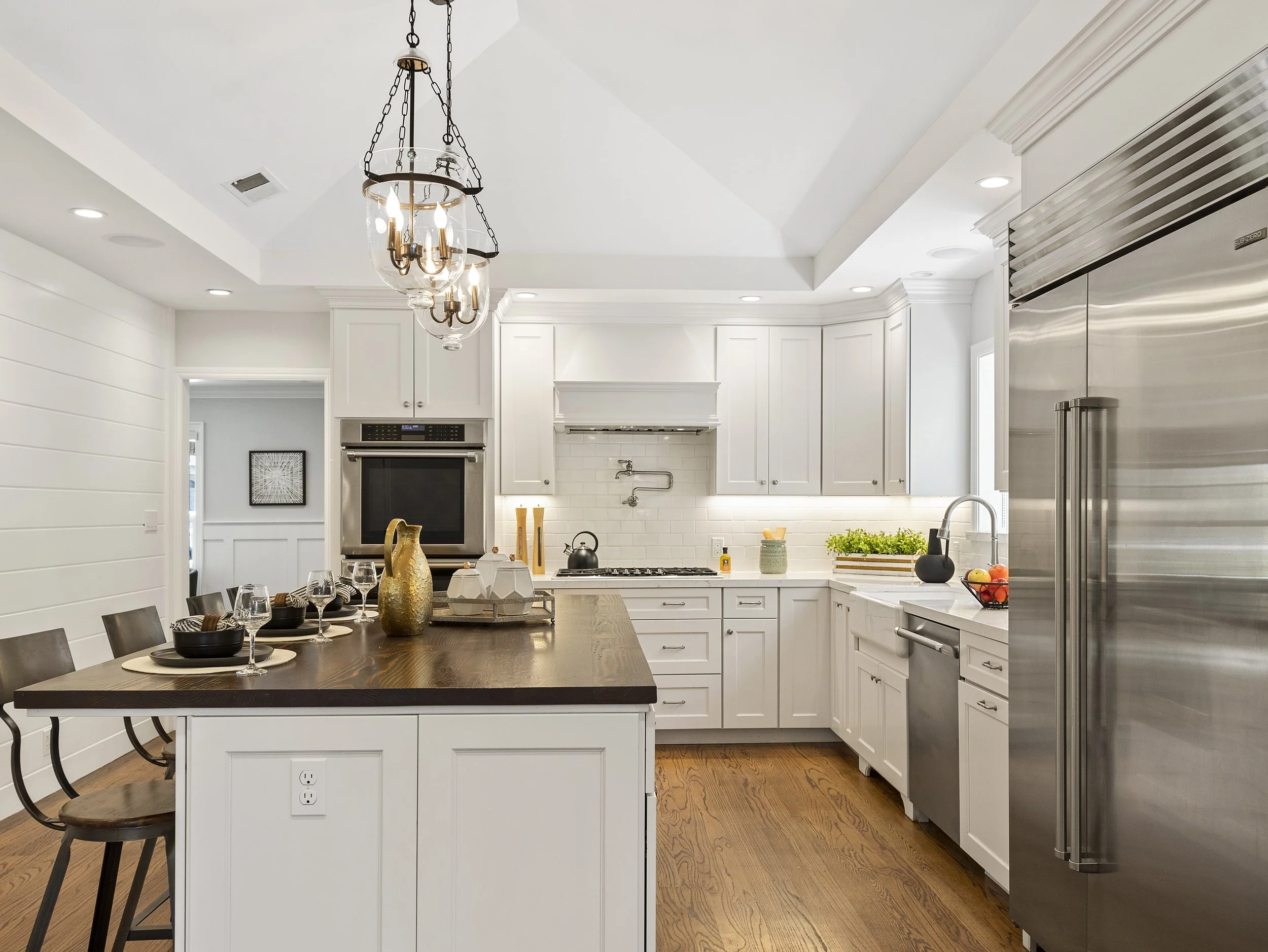 Modern kitchen with white cabinets, stainless steel appliances, and a dark wooden dining table with chairs. A chandelier hangs from the ceiling, and the kitchen includes a built-in oven, a large refrigerator, and a sink on the white countertop with d