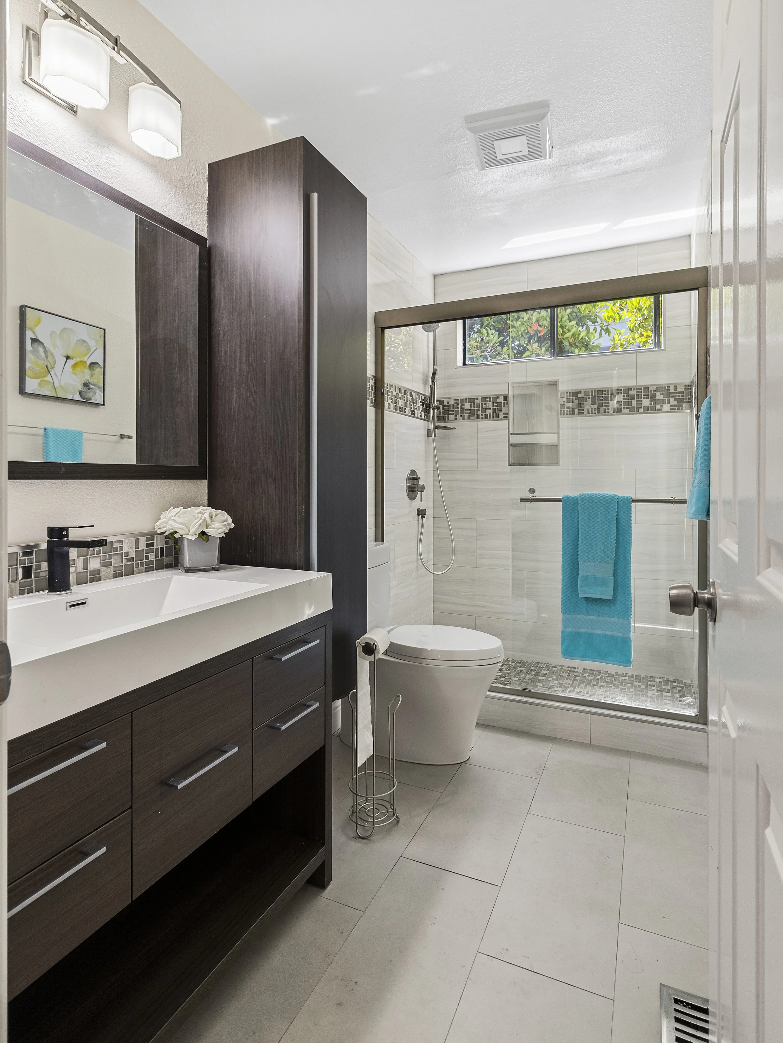 Modern bathroom with dark wood vanity, white sink, black faucet, large mirror, flowers in a white vase, shower with glass door, window with greenery, turquoise towel, and tiled walls.