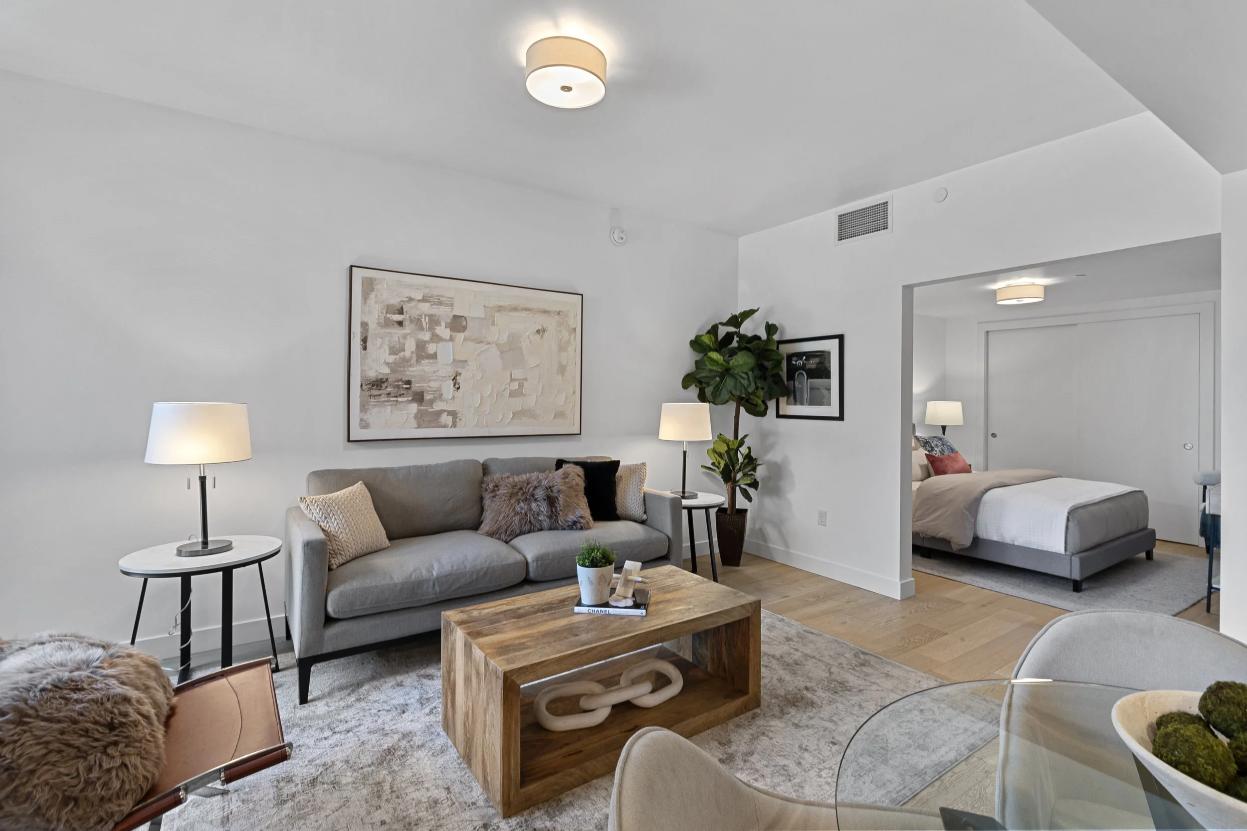 Living room with a beige sofa, two white lamps, wooden coffee table, abstract wall art, large plant, and a view into a bedroom with a bed and additional lamps.