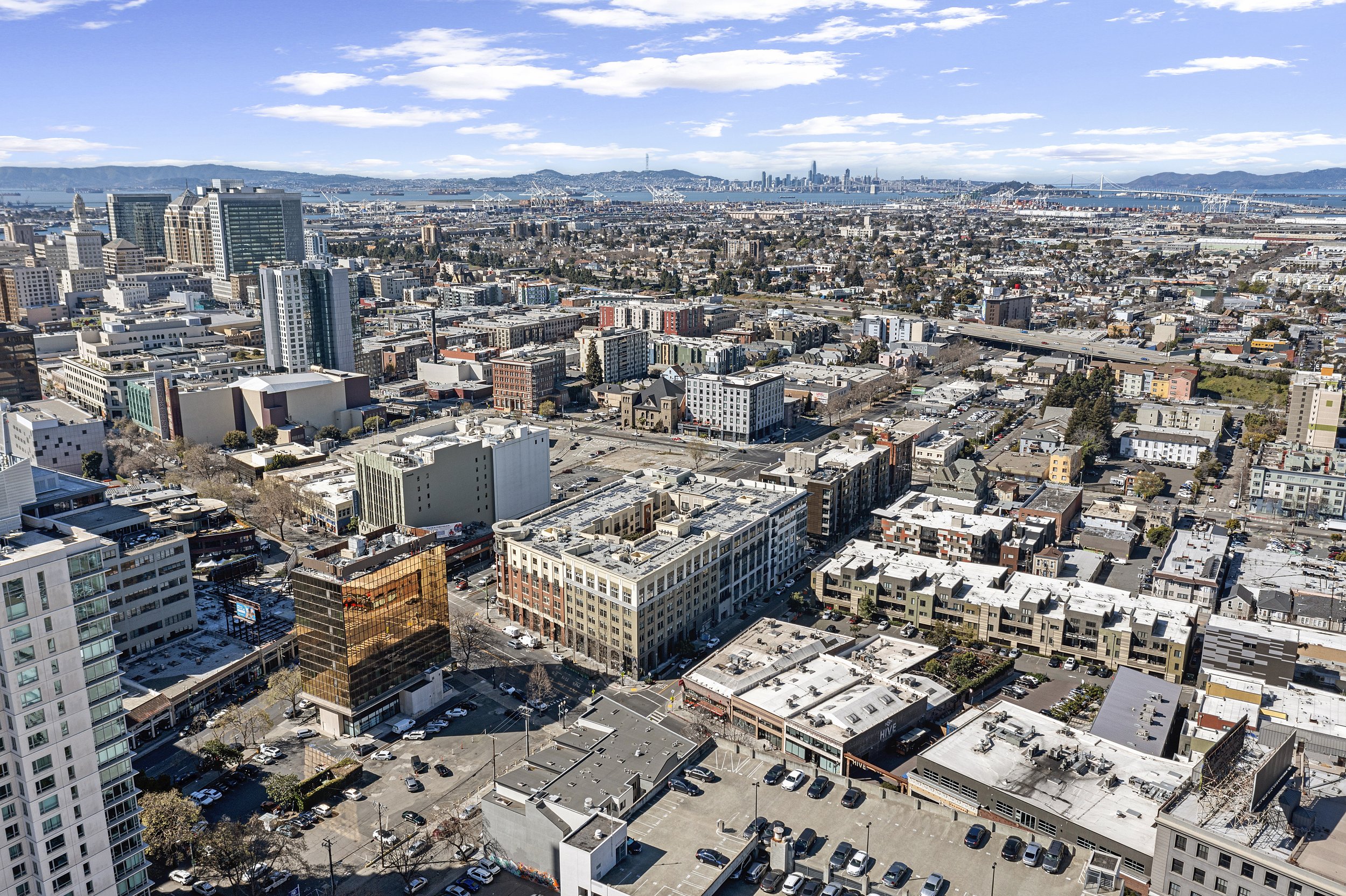 Aerial view of a cityscape with tall buildings, streets, and a bay with ships and a bridge in the background.