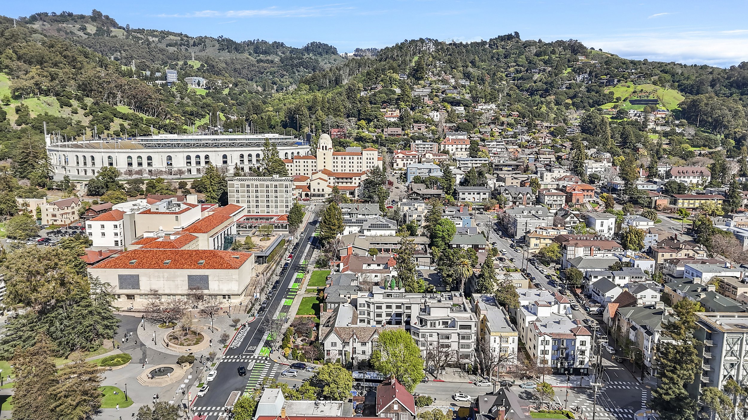 Aerial view of a city with a stadium, residential houses, and green hills in the background on a clear day.