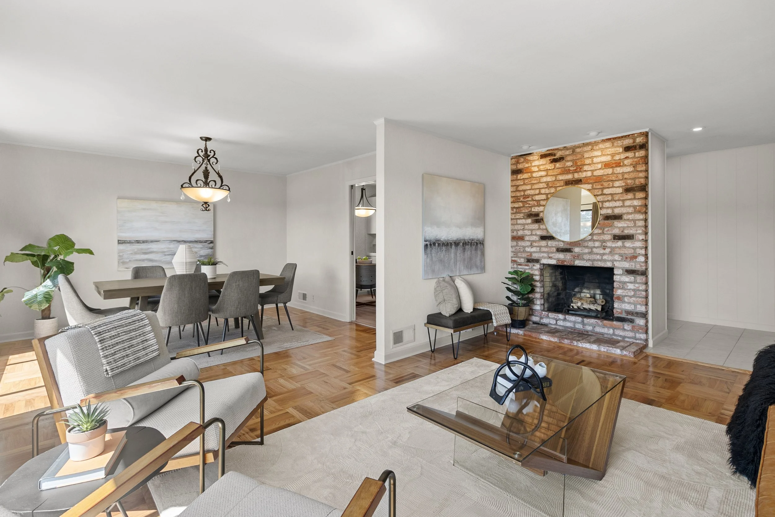 A spacious living and dining room with a brick fireplace, a round mirror, a dining table with chairs, and various indoor plants and artwork.