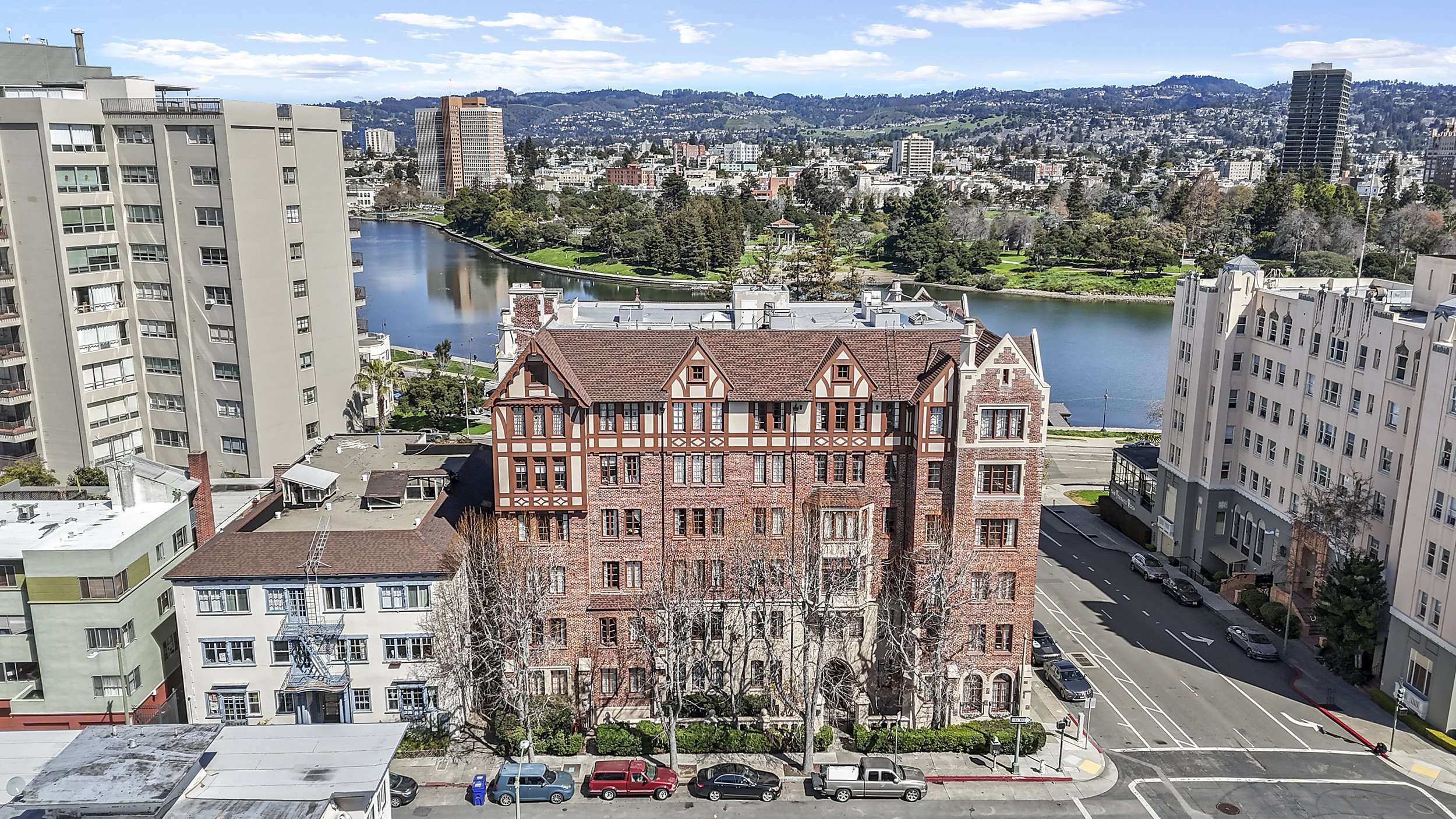 A cityscape featuring a river, tall apartment buildings, and a historic brick building with a gabled roof. Green hills and additional buildings are in the background under a partly cloudy sky.