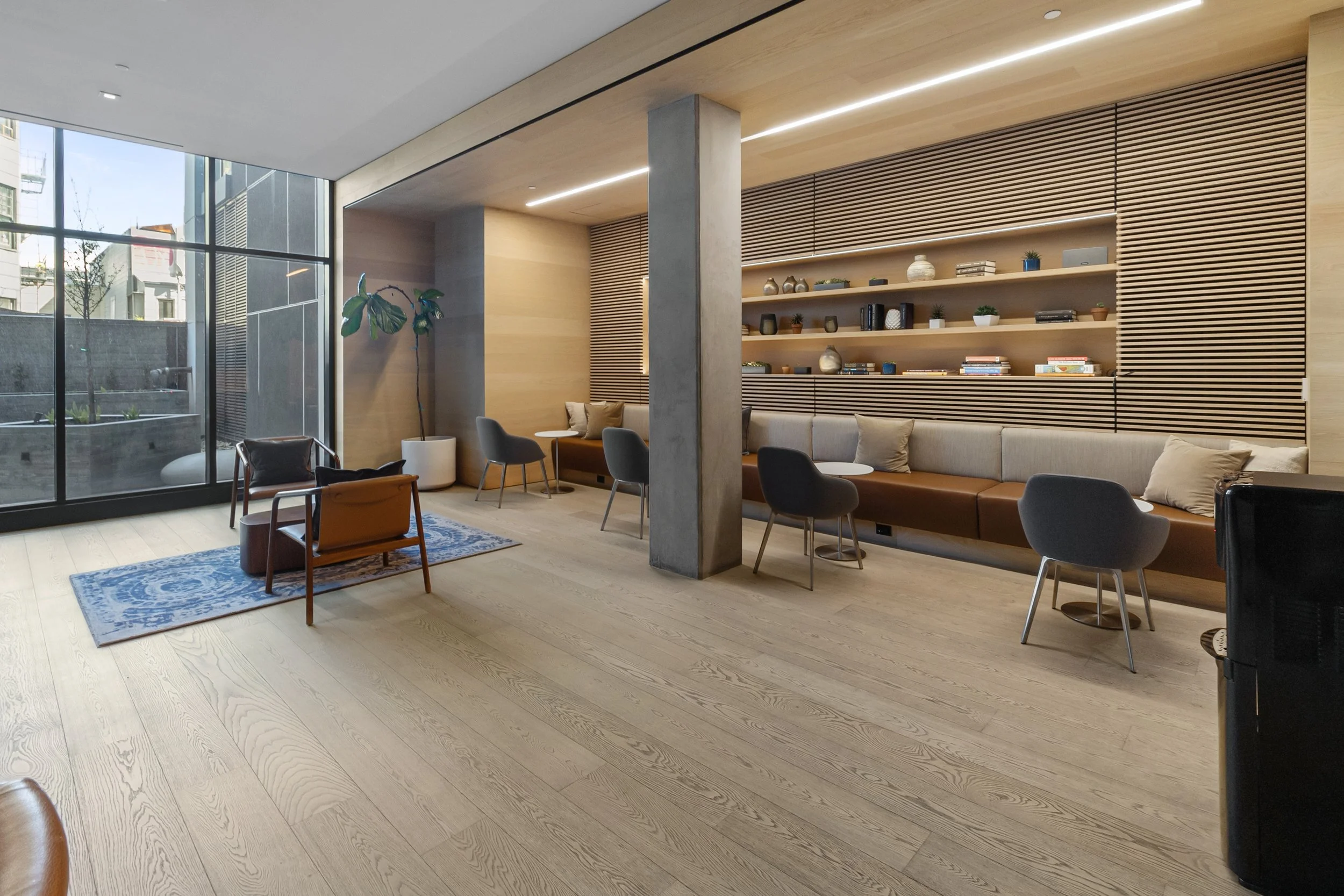 Modern lounge area with large floor-to-ceiling windows, wooden shelves with decorative items, gray chairs, beige sofa, potted plant, and light wood flooring.