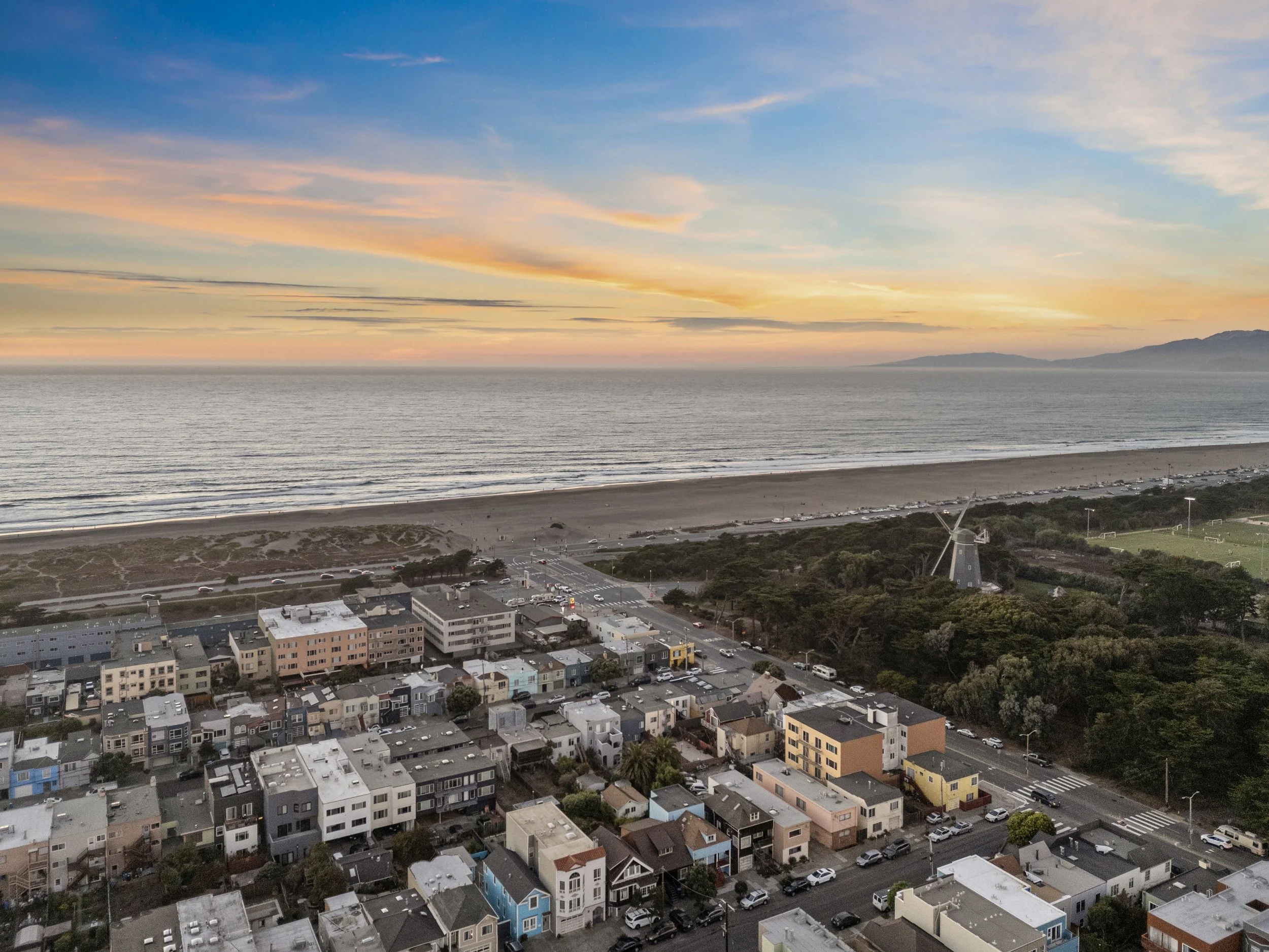 An aerial view of a coastal city at sunset, showing a beachfront, residential buildings, and streets surrounded by greenery with hills in the background.