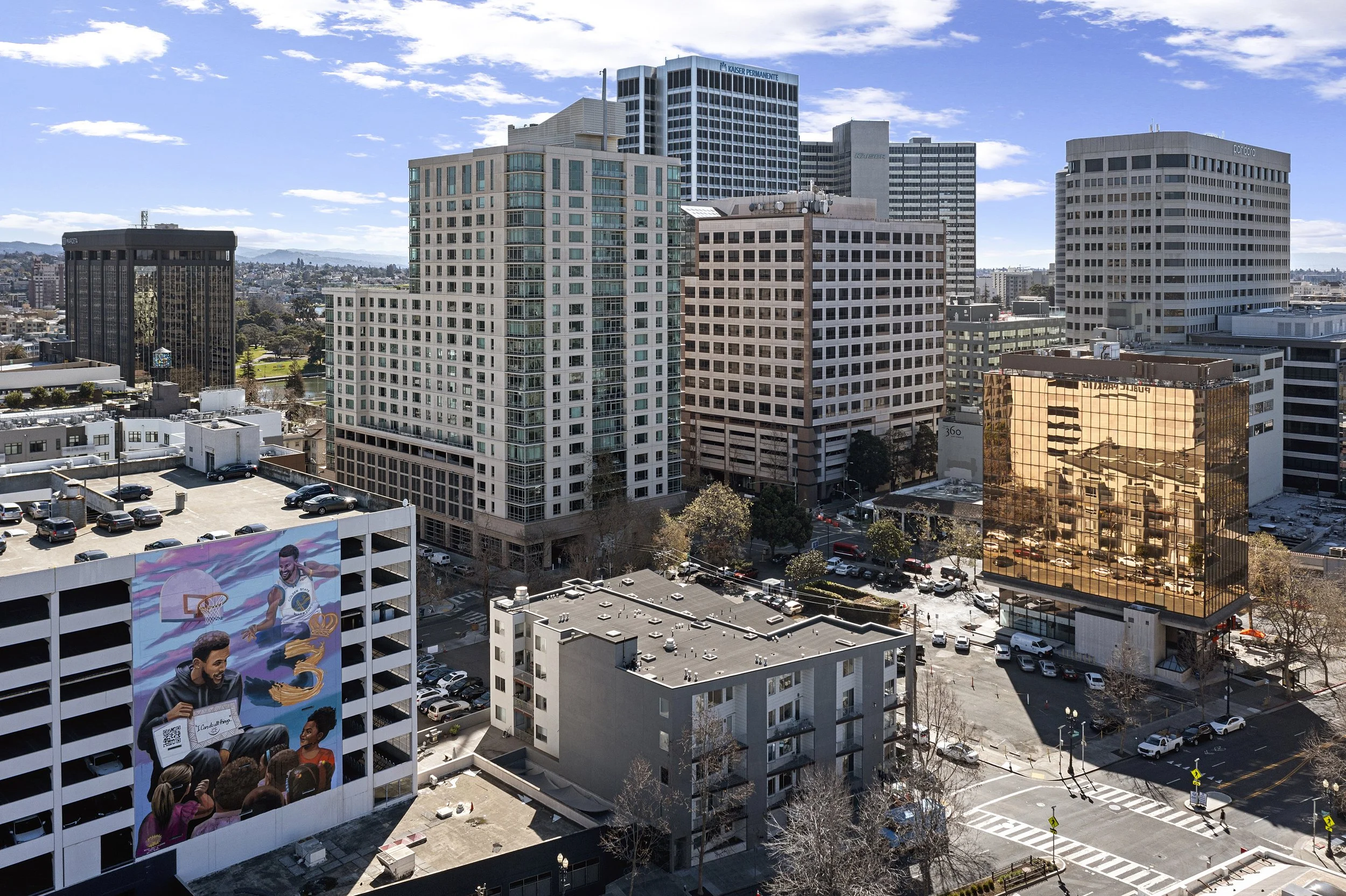 City skyline with tall modern buildings, some with glass facades reflecting the sky. A parking lot with cars is visible in the foreground. The sky is partly cloudy with patches of blue.