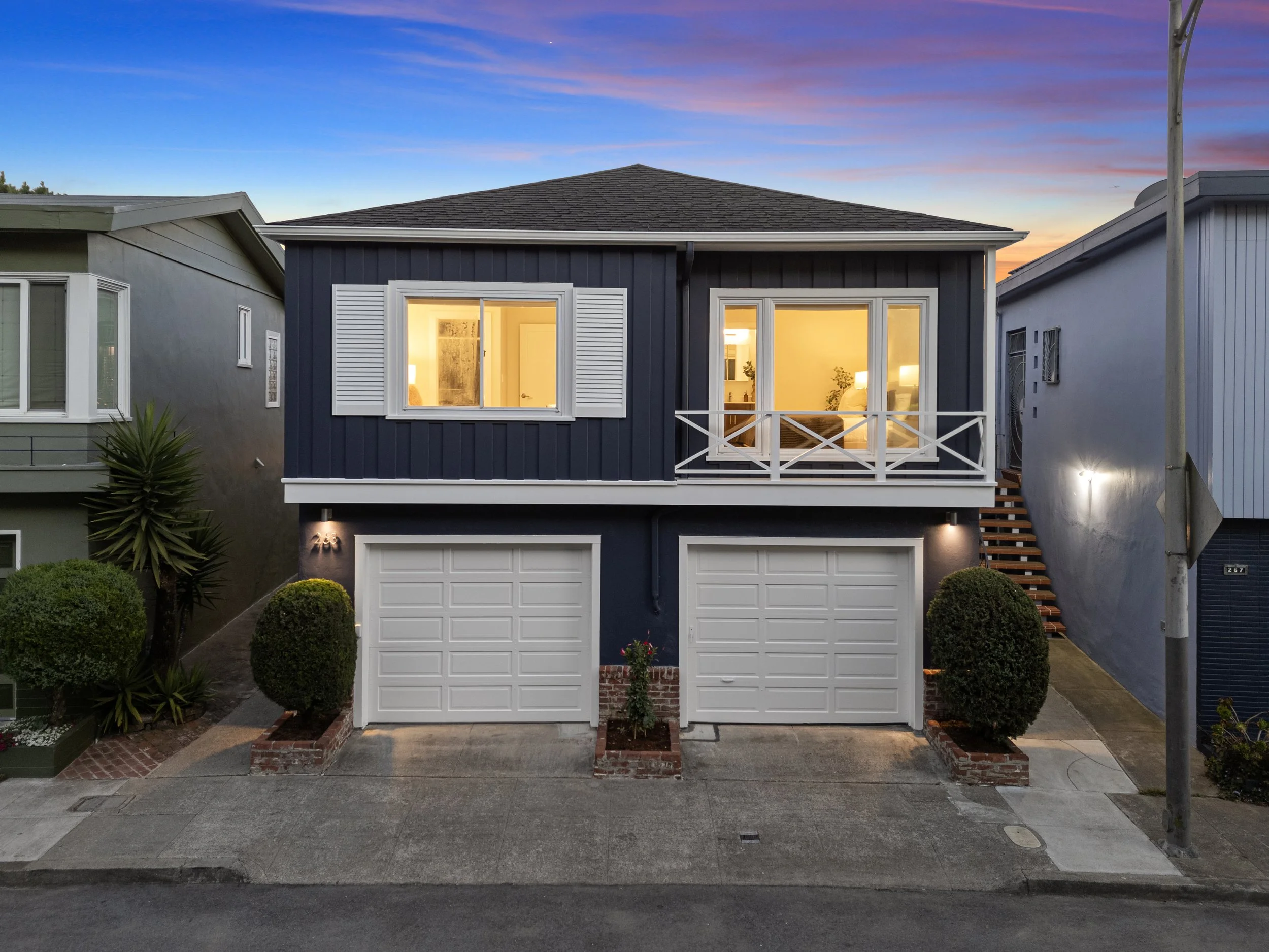 A two-story house painted dark blue with white trim, featuring two garage doors on the ground level, a balcony with a white railing on the second floor, and illuminated interior rooms visible through large windows. The house is flanked by similar nei