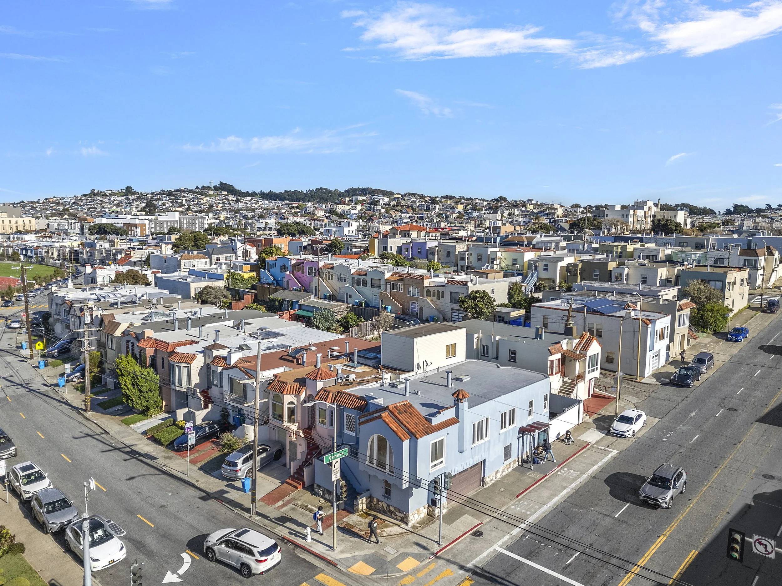 A neighborhood view of residential houses with colorful facades, parked cars along the streets, and a blue sky with scattered clouds.