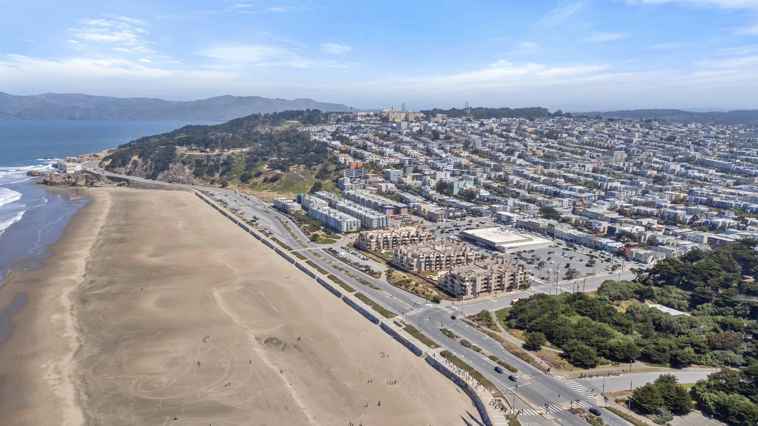 Aerial view of a coastal city with a beach, a road, and residential and commercial buildings with hills and mountains in the background under a partly cloudy sky.
