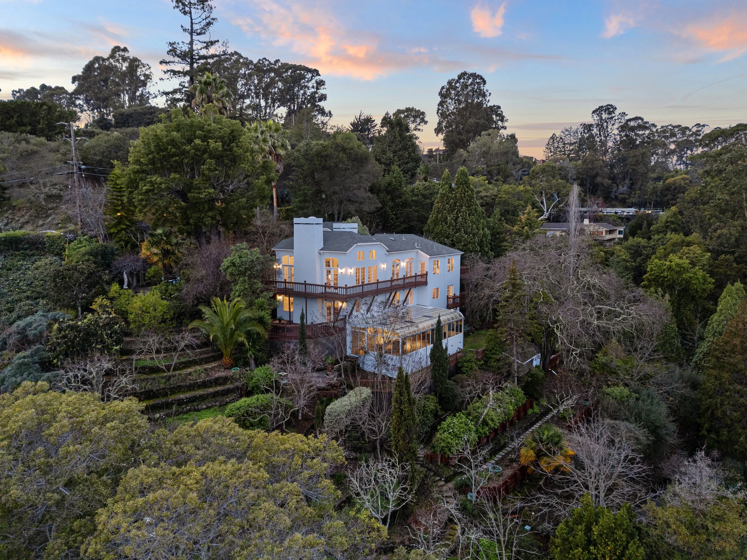 A house surrounded by a lush, green wooded hillside at sunset with a colorful sky and clouds in the background.