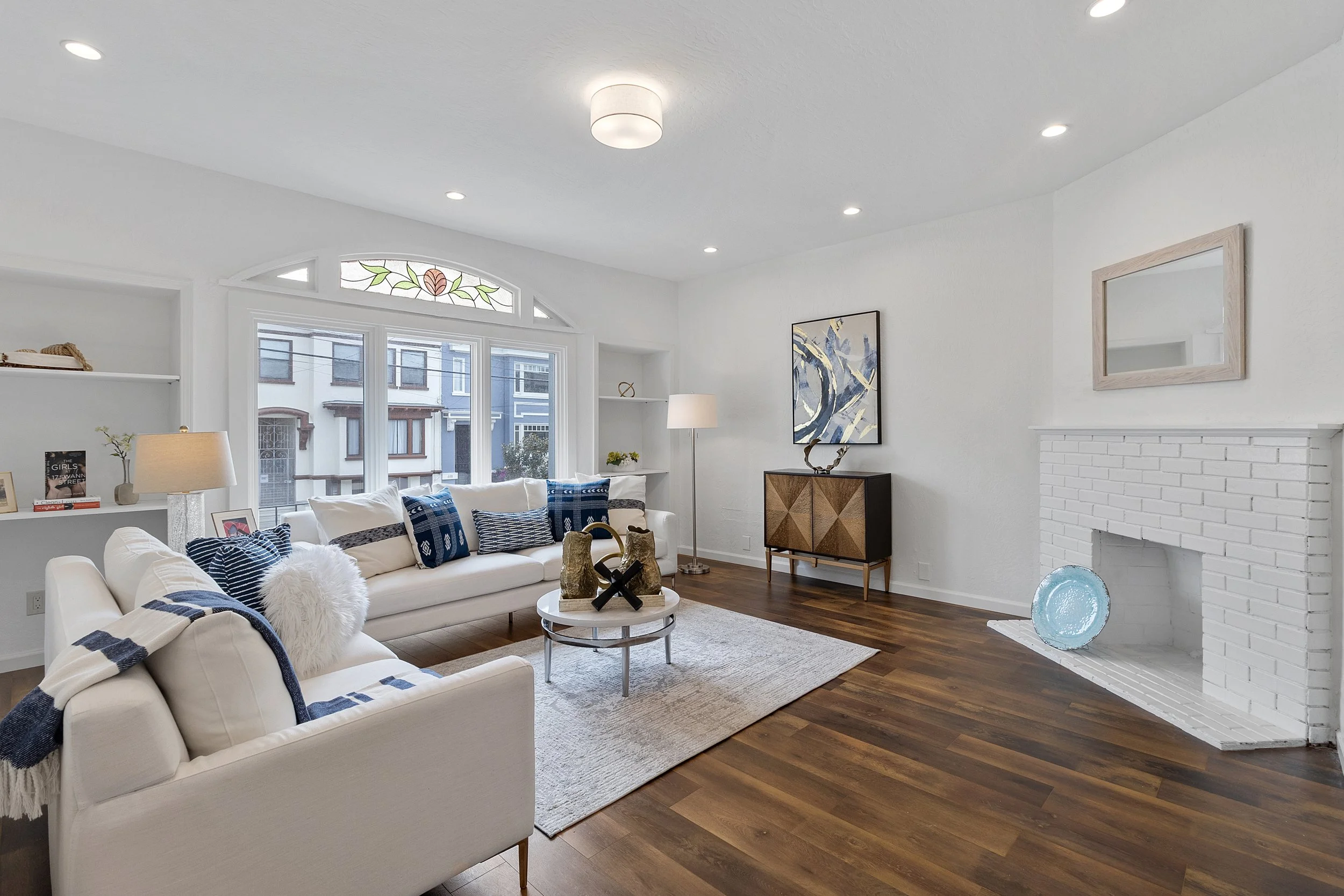 Living room with white sofa, blue pillows, wooden floor, white brick fireplace, artwork, and large window with stained glass detail.