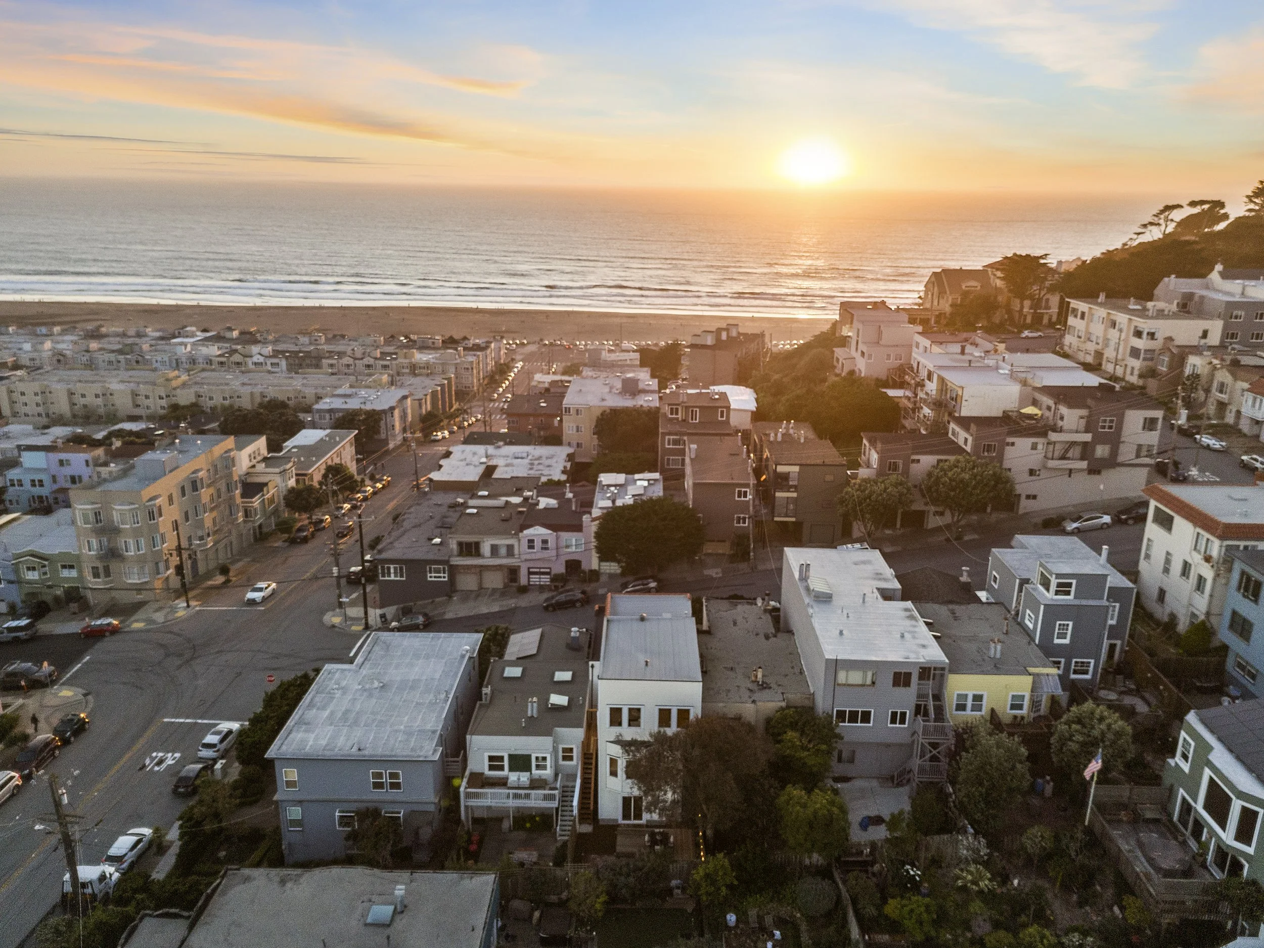Aerial view of a coastal town during sunset with residential buildings, a beach, and the ocean in the background.