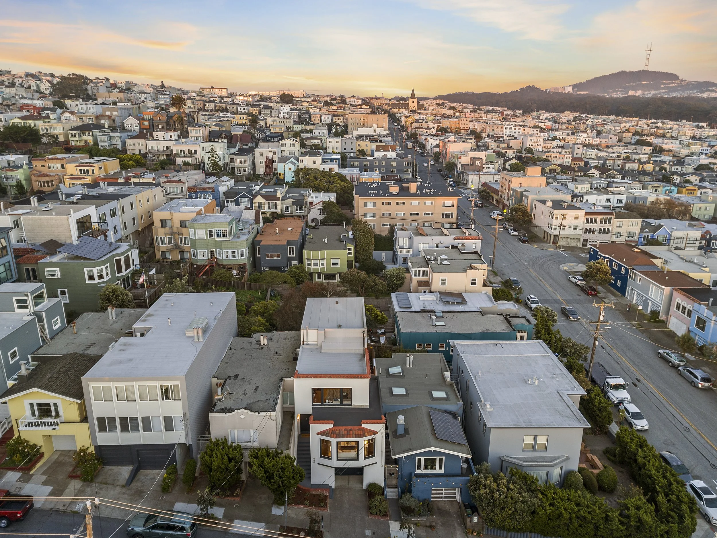 Aerial view of a neighborhood with colorful houses, streets, cars, and a church steeple, with hills in the background during sunset.