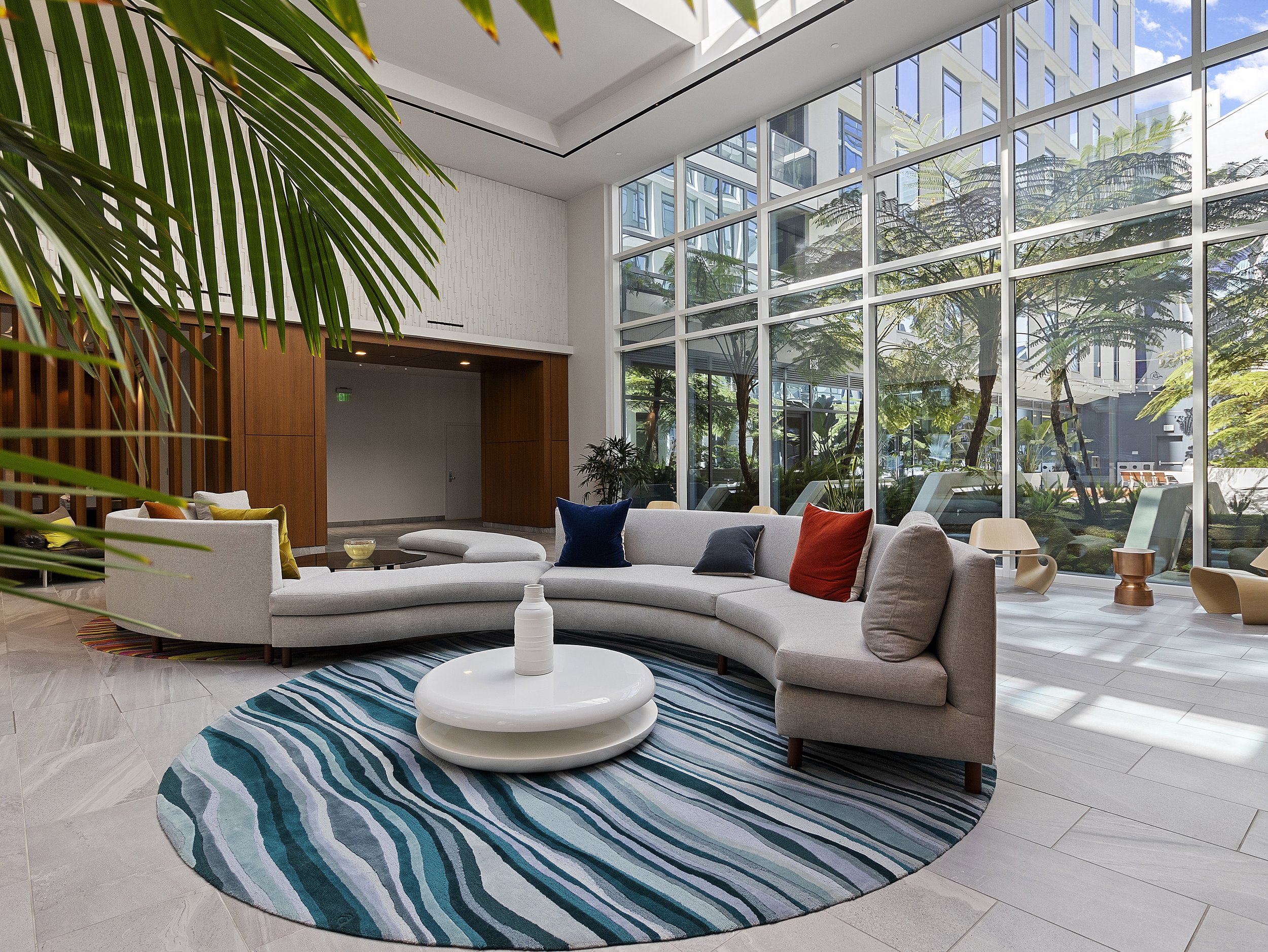Modern hotel lobby with large floor-to-ceiling windows, white curved sofa with colorful pillows, a round white coffee table with a white vase, a blue and green striped rug, indoor plants, and high-rise buildings visible through the windows.