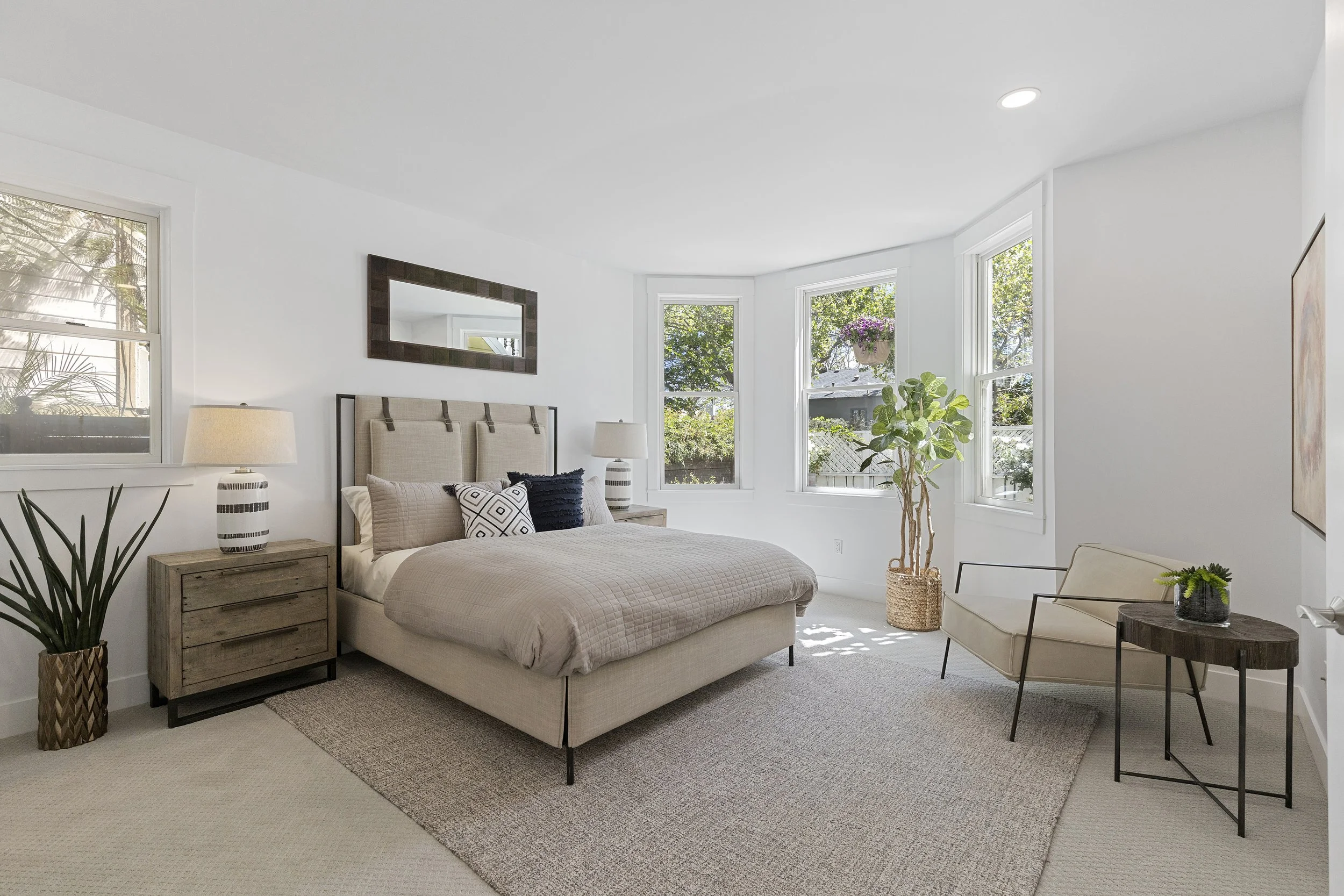 Bright bedroom with large windows, beige bed with decorative pillows, wooden nightstand, lamps, and indoor plants.