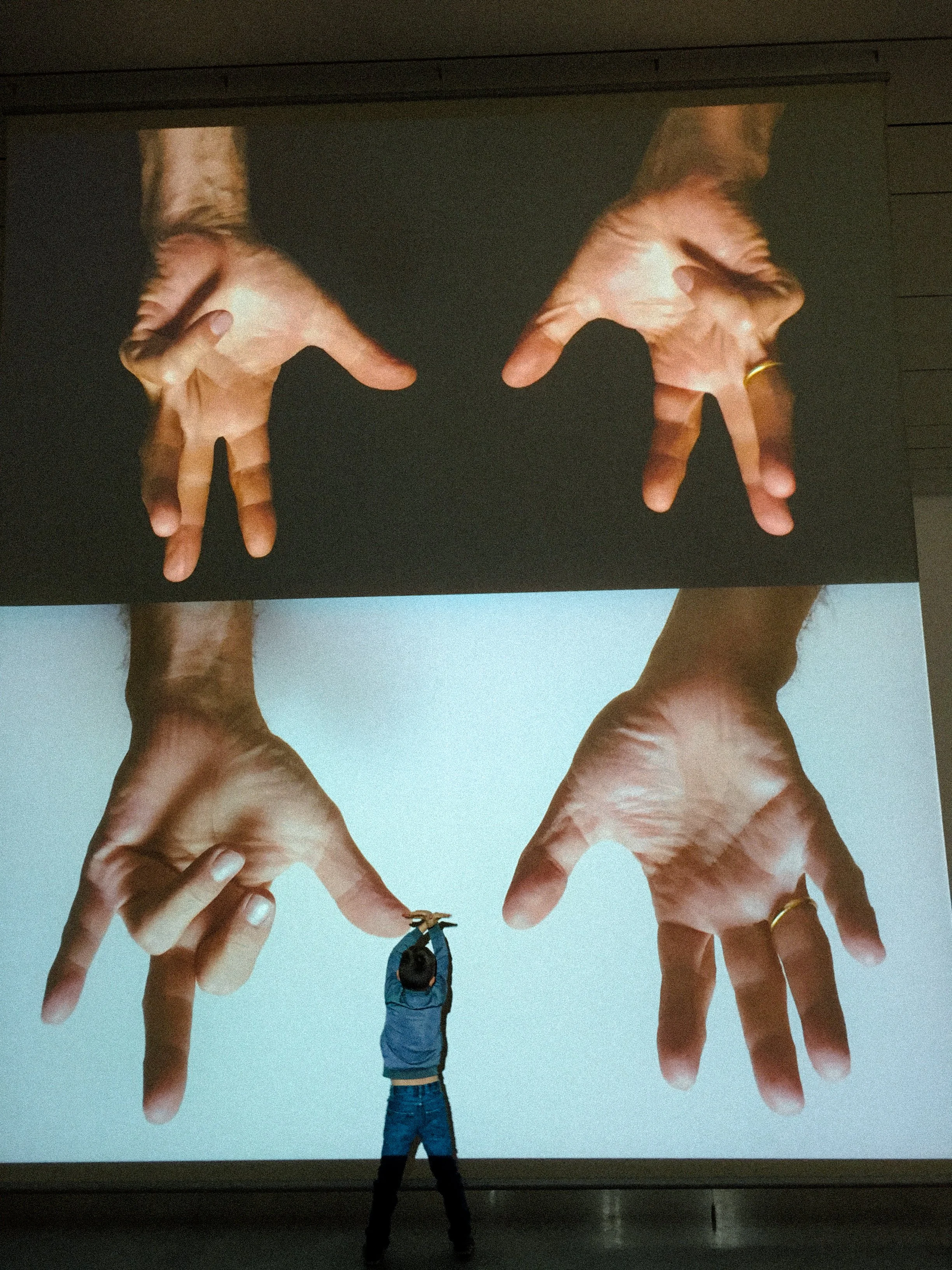 A person in a blue jacket and jeans stands with arms raised in front of a large screen showing four oversized hands making various gestures, with the person’s head near the middle of the hands.