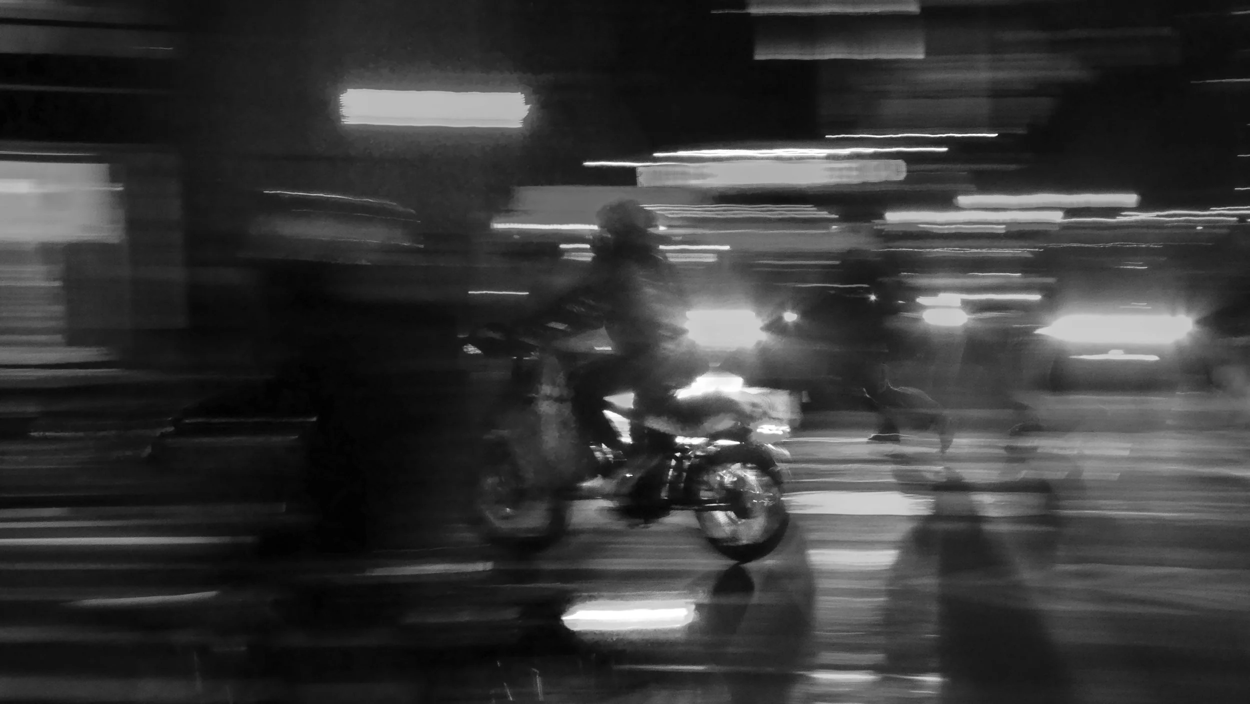Blurry black and white photo of a person riding a motorcycle at night, with streaks of lights from cars and streetlights in the background.