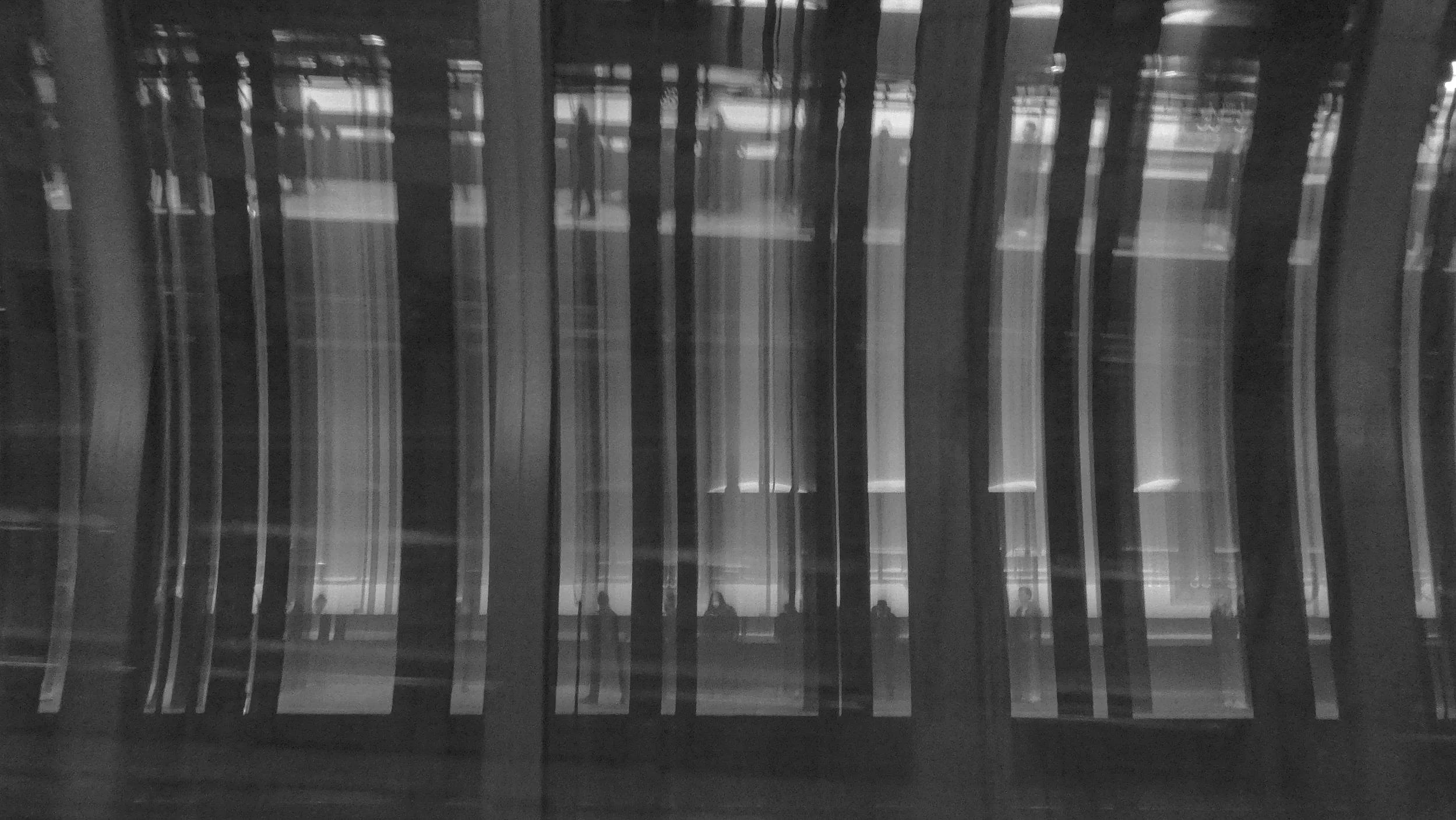 Interior view of a modern building with tall glass walls, reflecting silhouette figures of people inside.