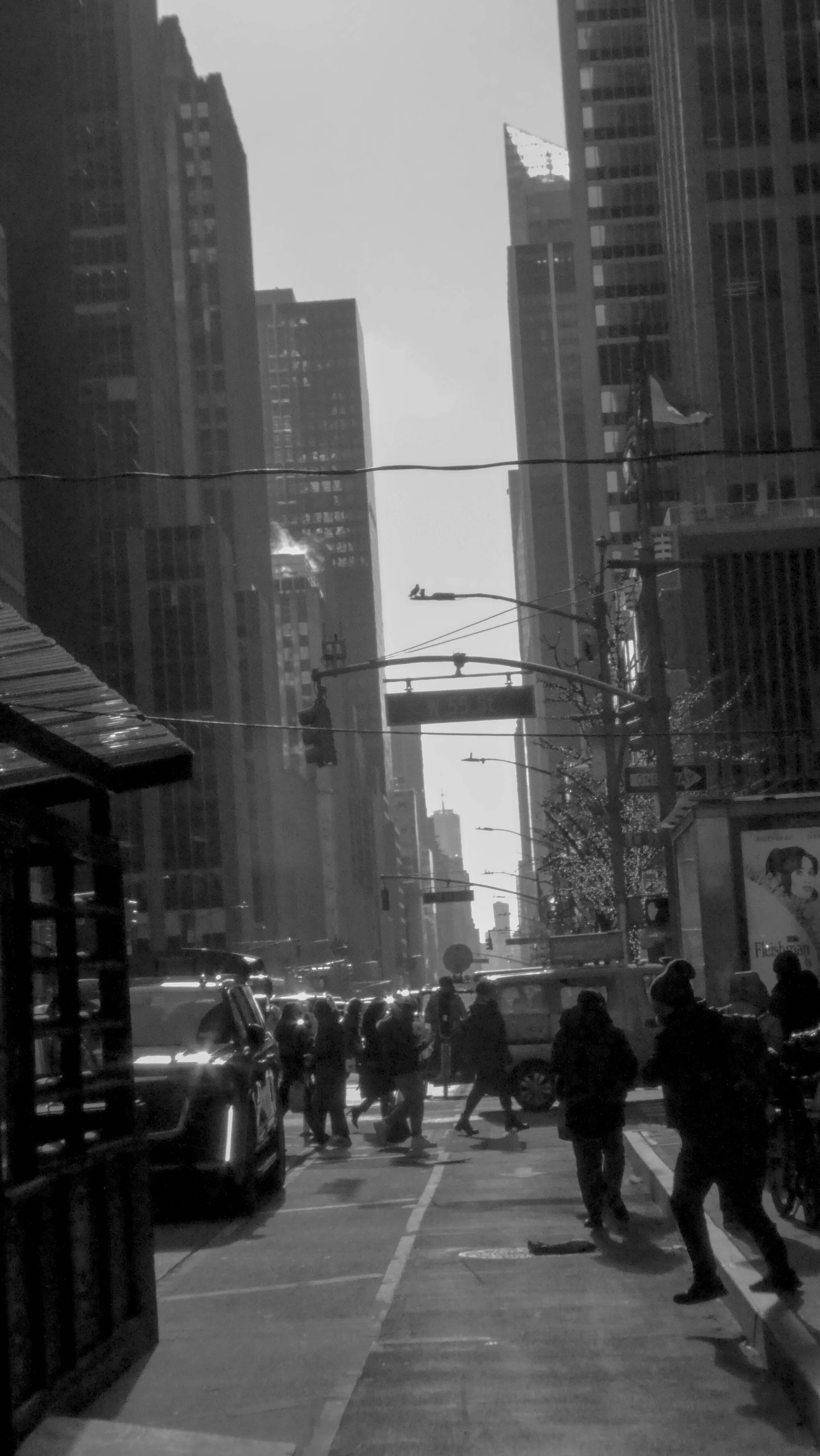Black and white photo of a busy city street with pedestrians crossing at a crosswalk, tall skyscrapers, and streetlights lining the sidewalk.