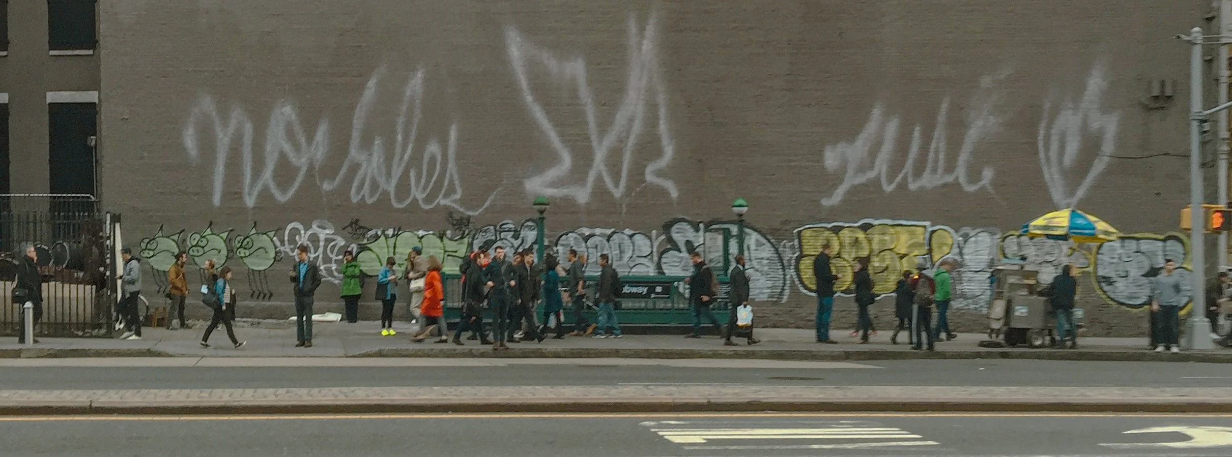 People walking along a city sidewalk in front of a wall covered in graffiti and large white chalk writing.
