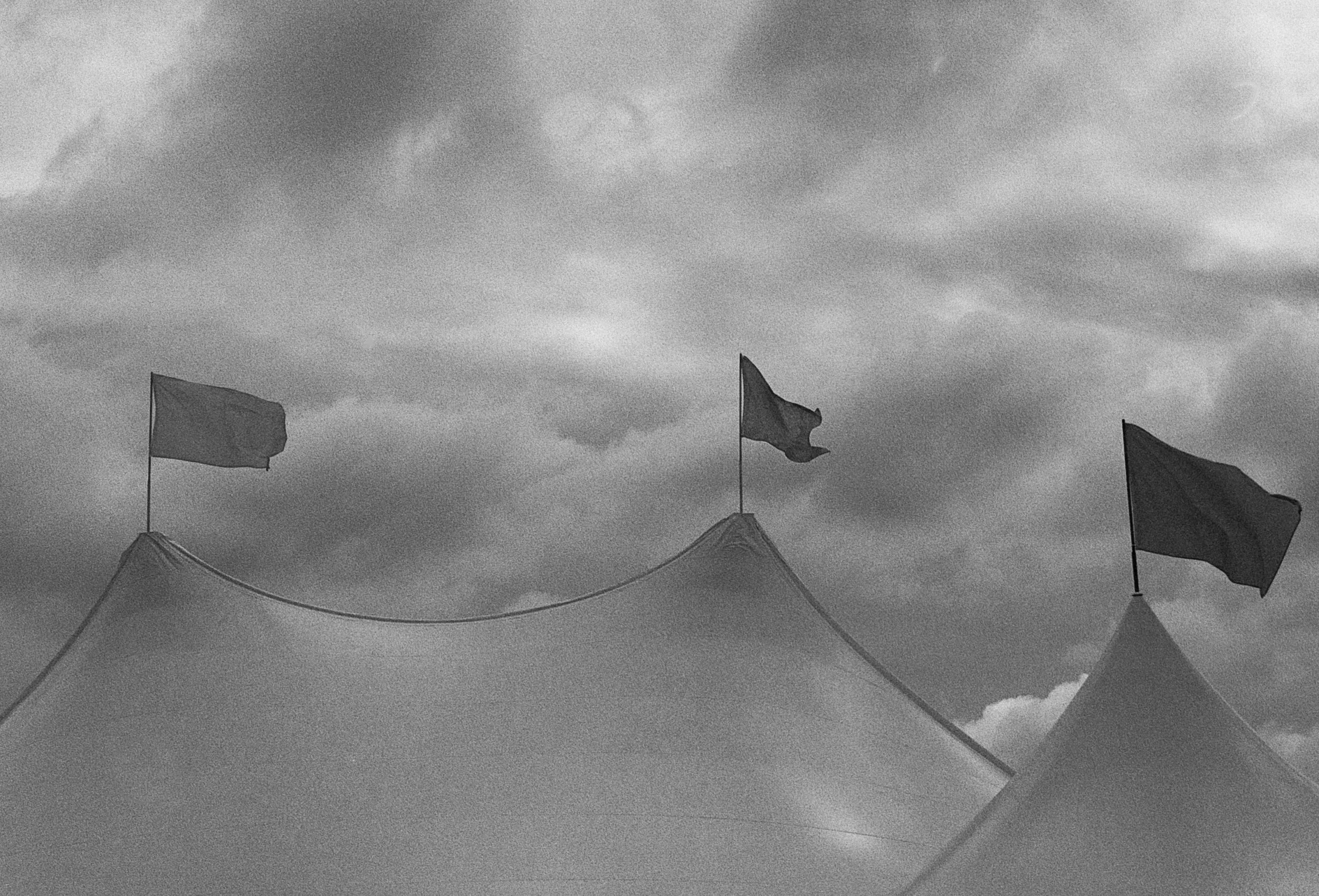 Three flags on poles atop tents against cloudy sky, black and white photo.