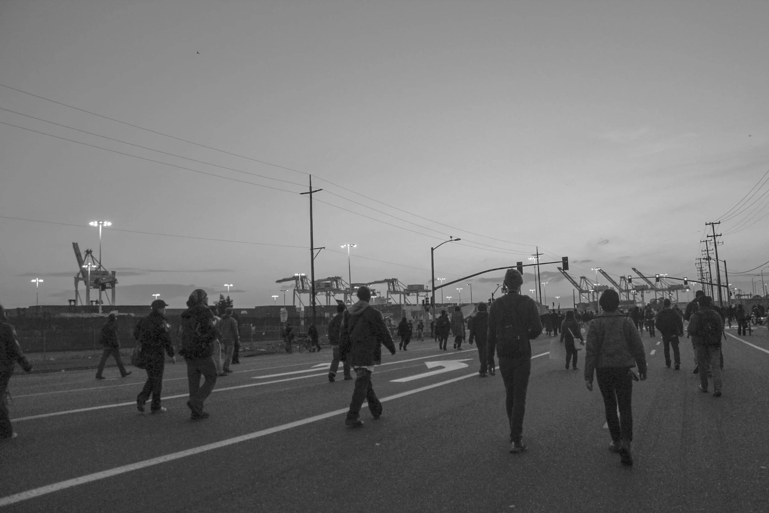 Crowd of people walking on a road near industrial cranes and power lines at dusk or dawn in an urban setting.