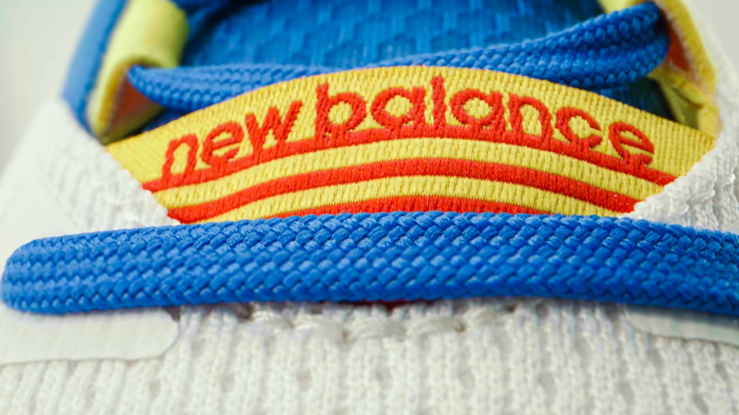 Close-up of a pair of white sneakers with blue laces, showing a yellow and red striped tag sewn on the tongue.