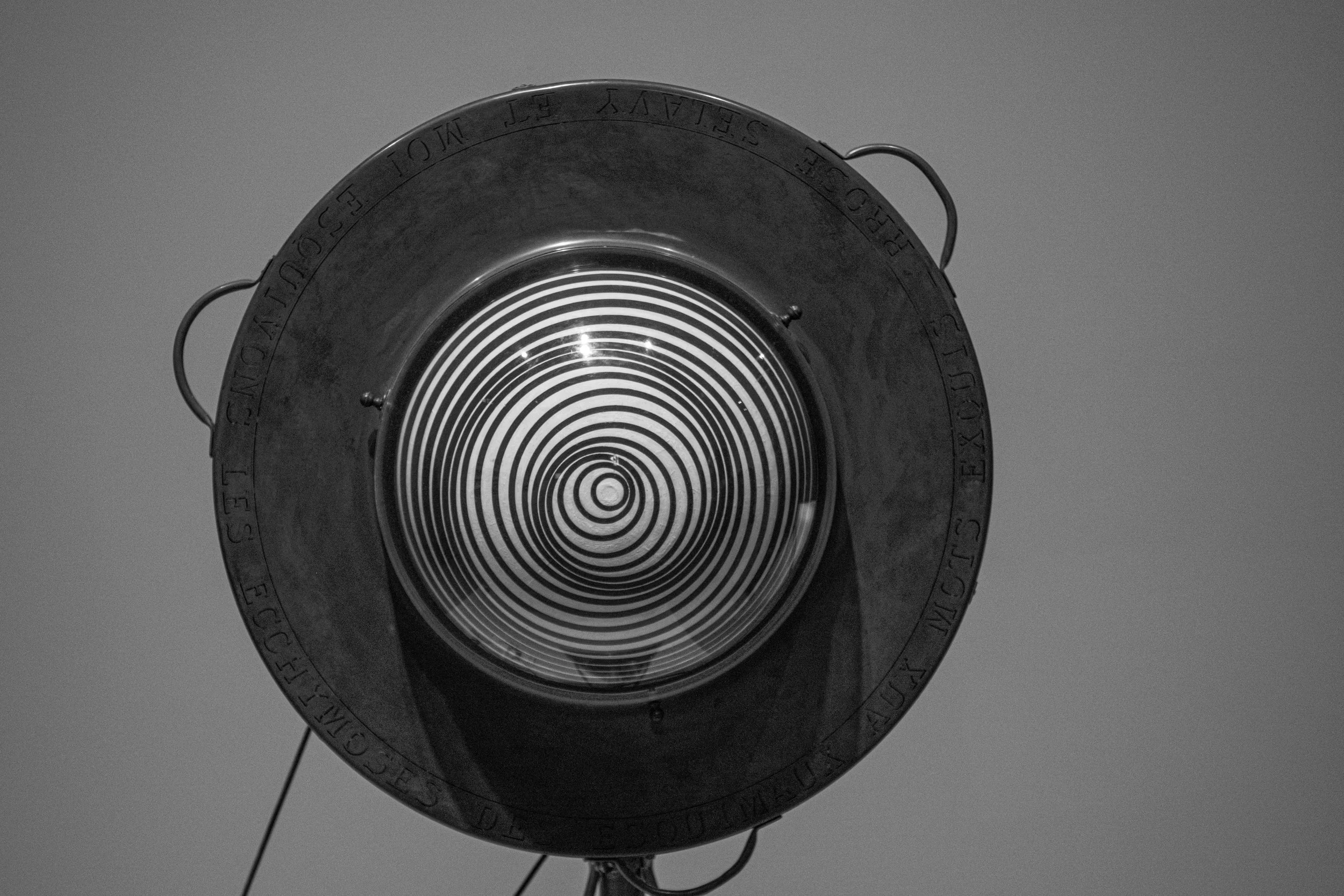Close-up of a vintage searchlight with a spiral-patterned lens, mounted on a stand against a plain sky