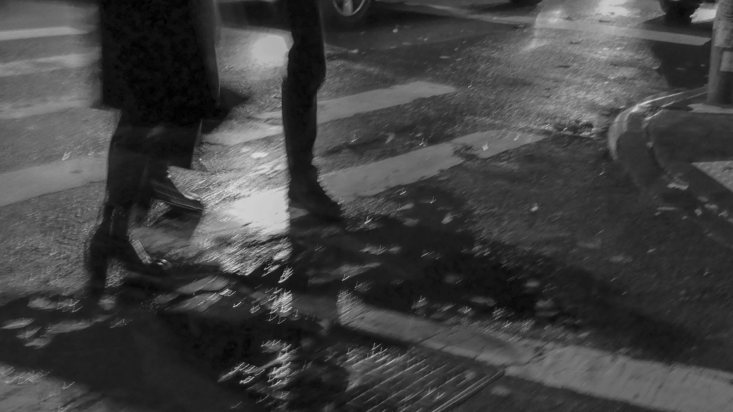 A person walking on a wet city street at night, wearing dark clothing and shoes, with their shadow visible on the ground.