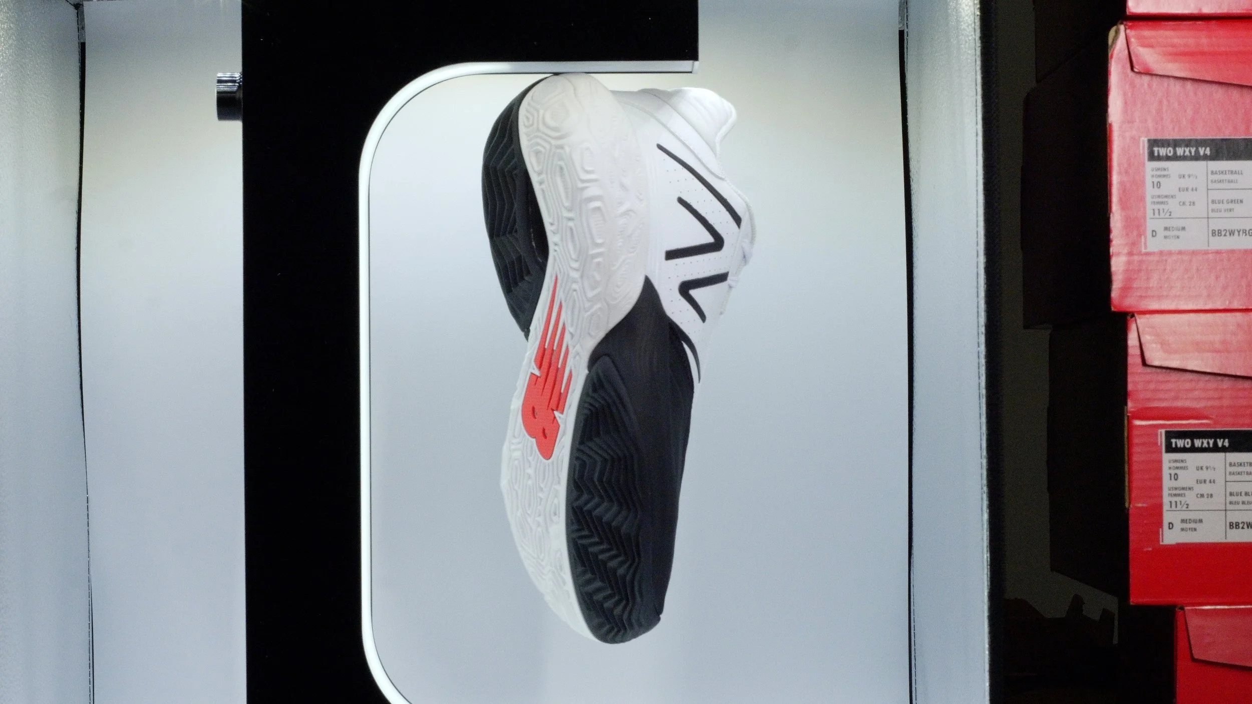 A white and black athletic shoe with red accents displayed on a lightbox at a store. The shoe is viewed from the bottom, showing the sole, and there are stacked red shoeboxes to the right.