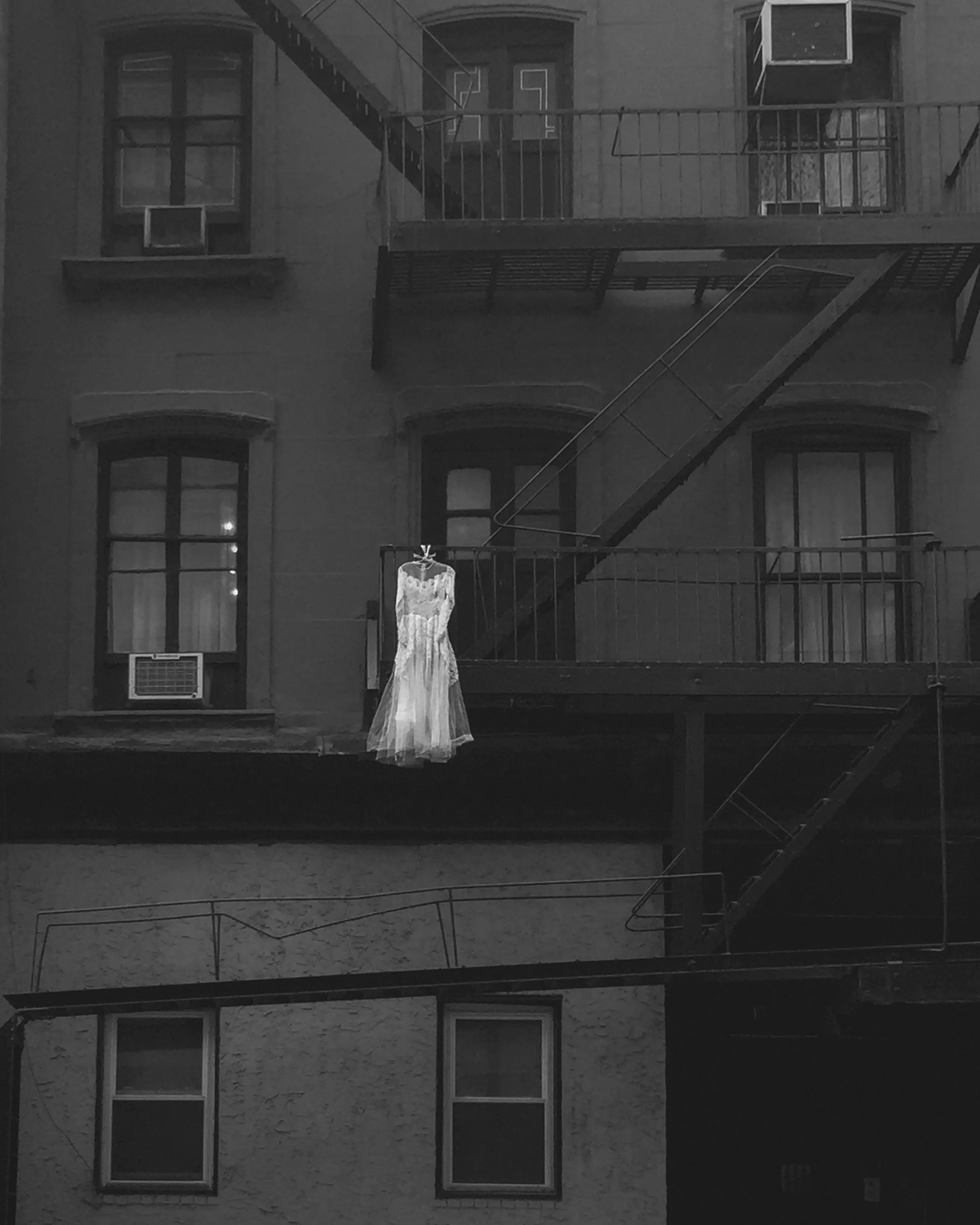 A white dress hangs on a hook on a fire escape outside an apartment building at night, with windows and air conditioning units visible in the background.