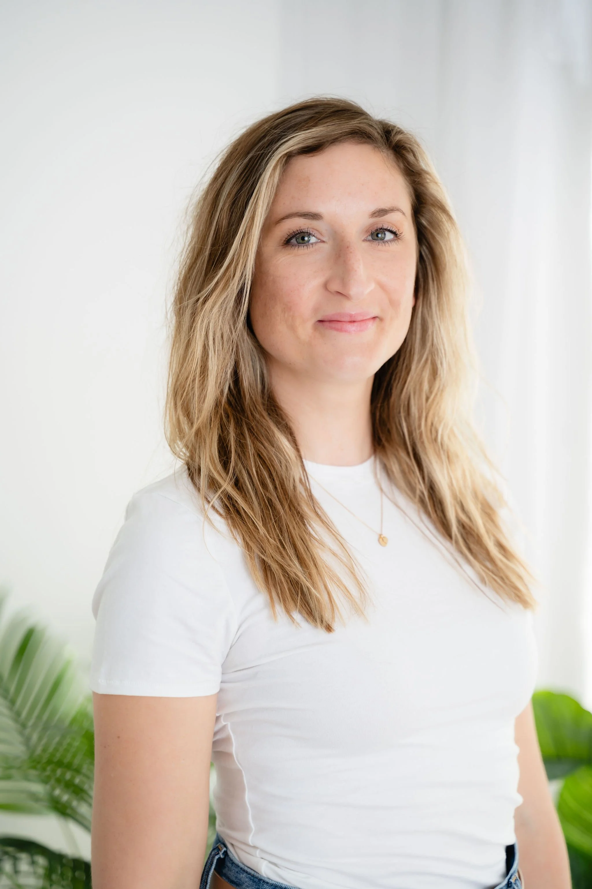 Close-up of a young woman with long, wavy, light brown hair and blue eyes wearing a white t-shirt, standing in front of a plain light background with some greenery visible.