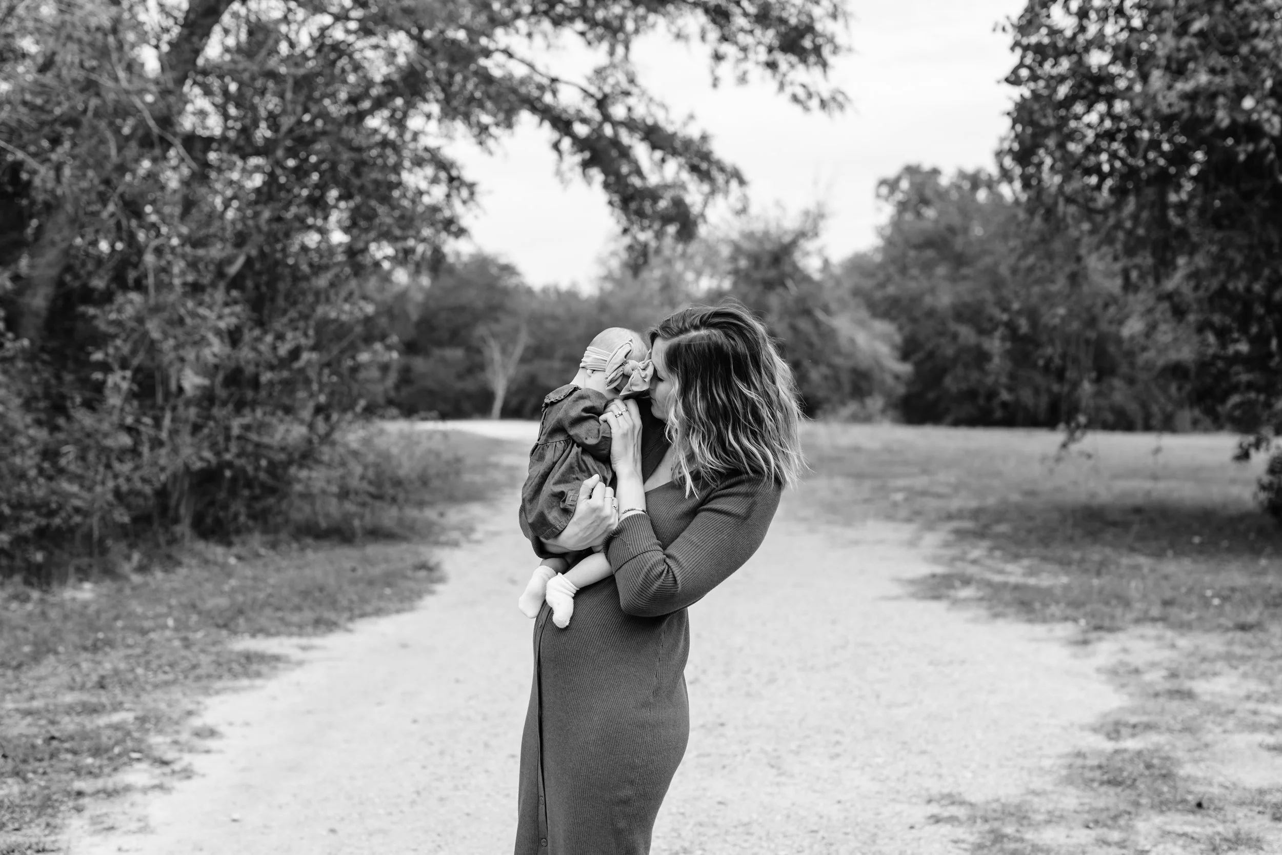 A woman holding a small child in an outdoor setting, with trees and a pathway in the background, in black and white.