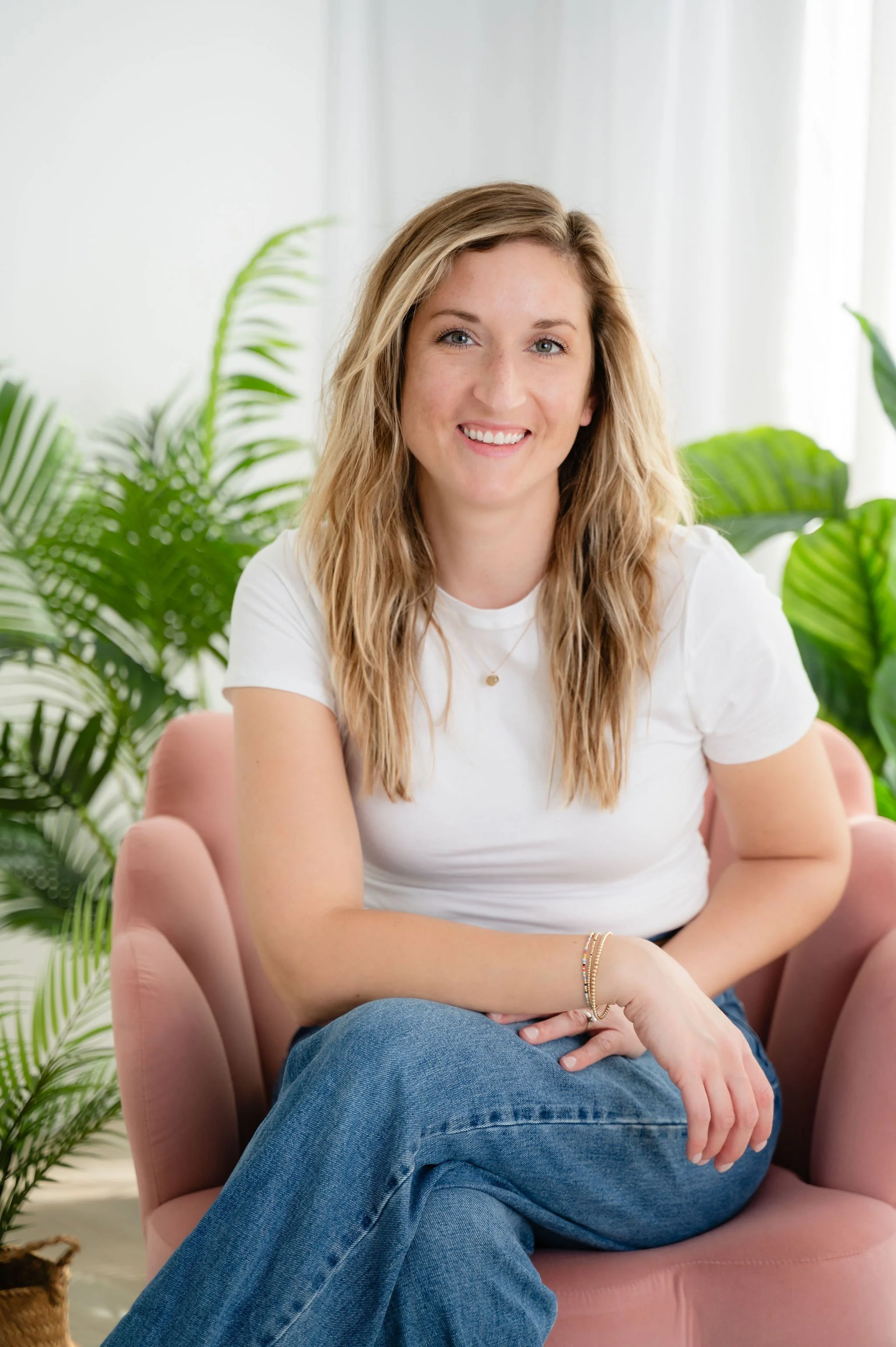 A woman with blonde hair and a white shirt smiling while sitting on a pink armchair surrounded by green plants.