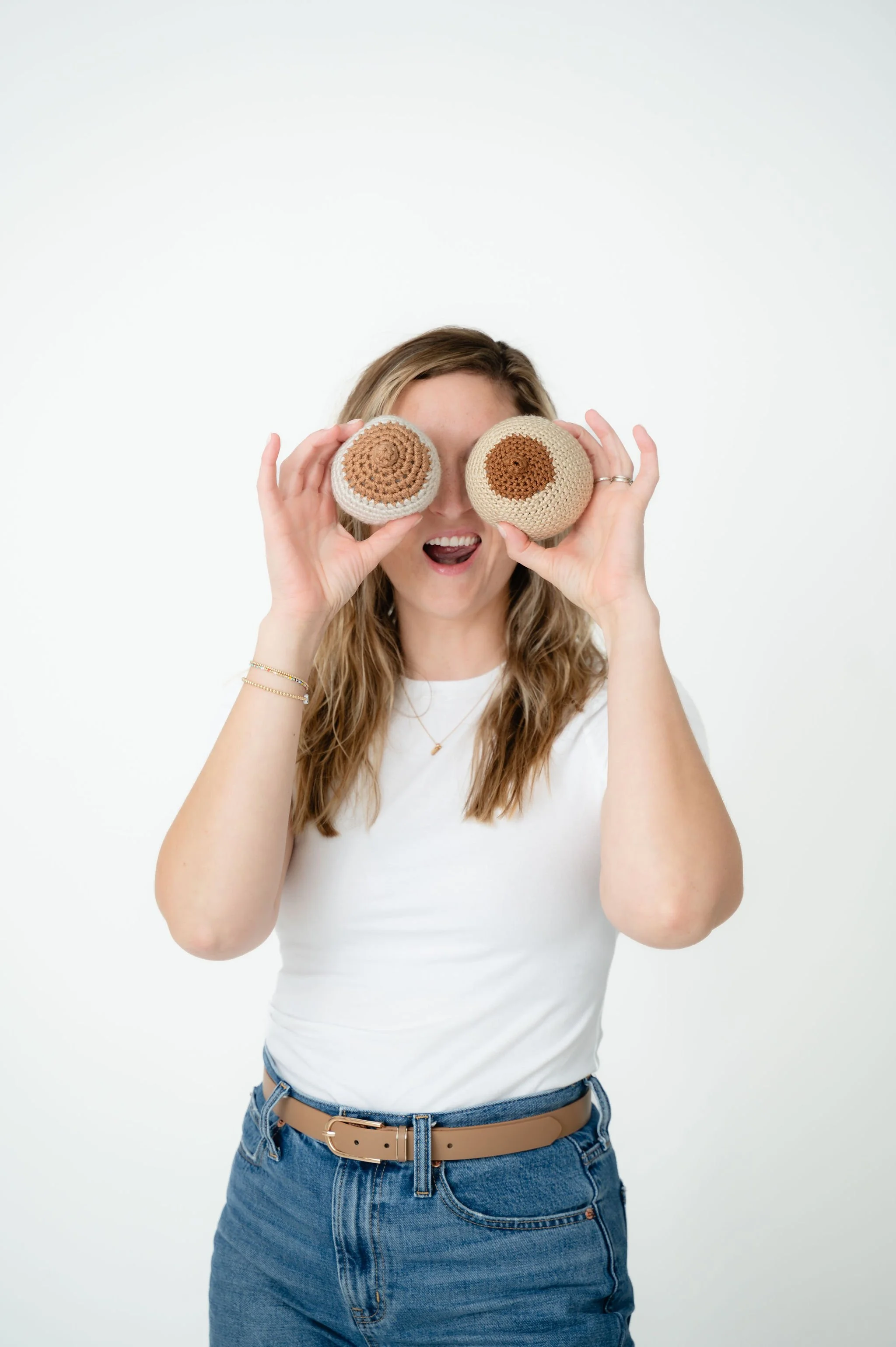 A woman wearing a white t-shirt and blue jeans is holding crocheted objects over her eyes, with her mouth open in an expression of surprise or delight.