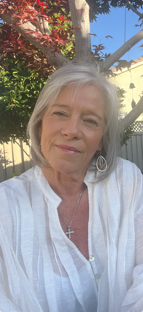 A woman with short gray hair smiling outdoors, wearing a white blouse and a cross necklace, standing in front of a tree with red leaves and a fence.