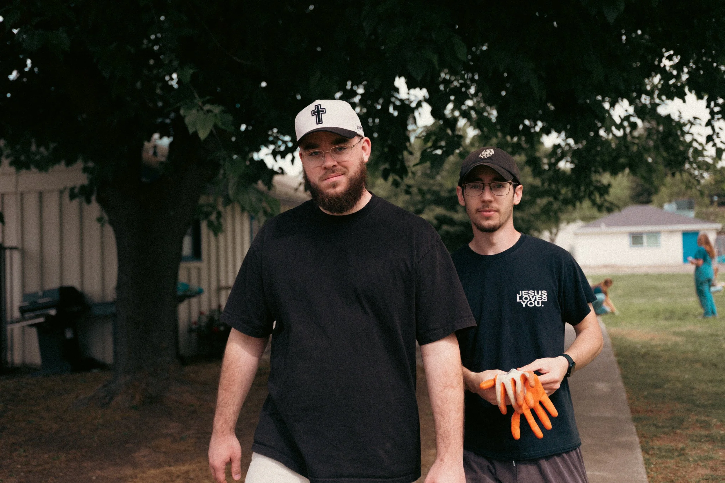 Two young men standing outdoors under a tree, one wearing a white cap with a cross symbol, glasses, and a black shirt, and the other wearing a black cap, glasses, a black shirt with white text, and holding orange gloves, with a building and other people in the background.