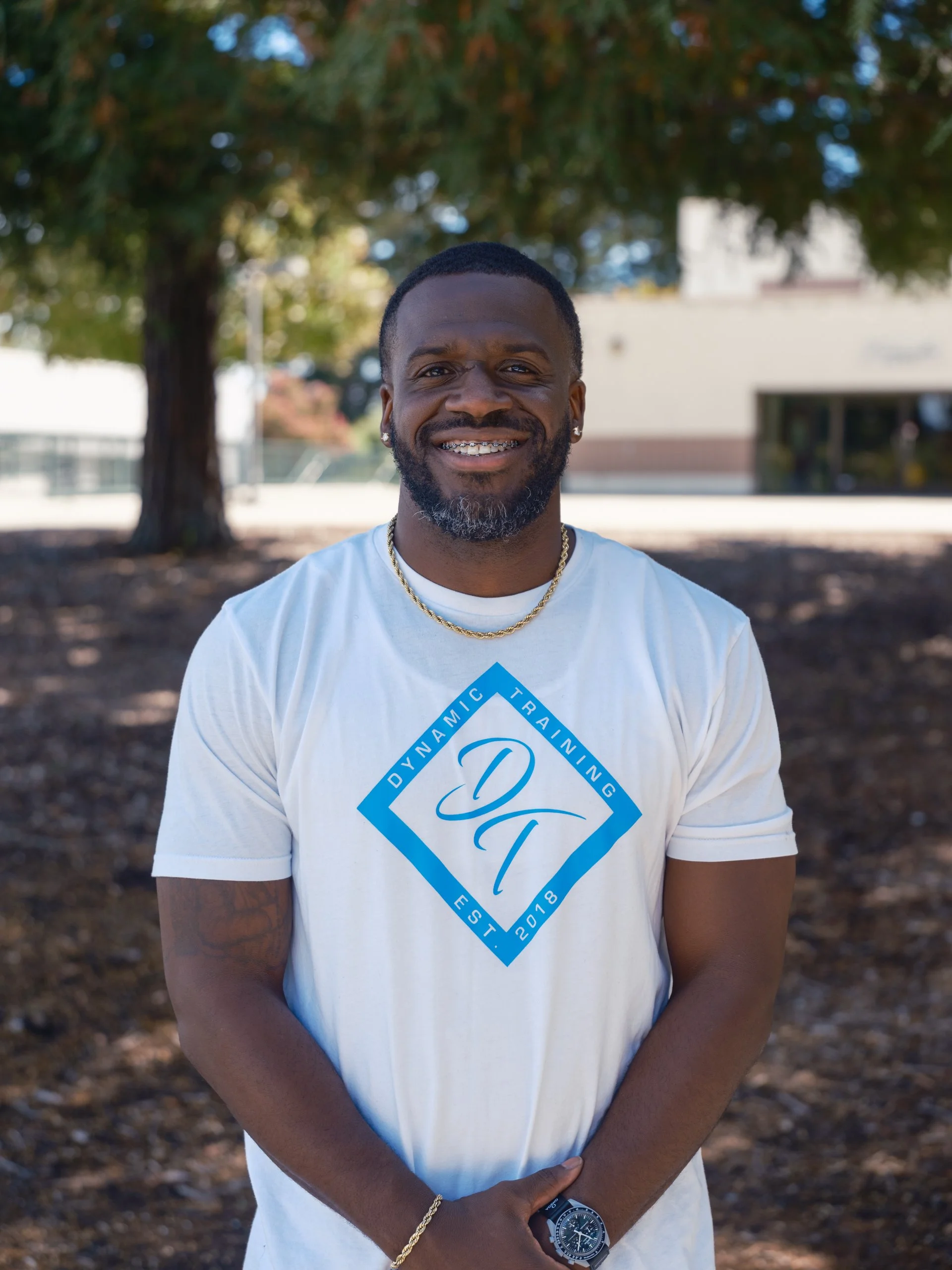 A smiling man with a beard and braces, wearing a white T-shirt with a blue logo, a gold chain, a black wristwatch, and earrings, standing outdoors under a tree with blurred buildings in the background.