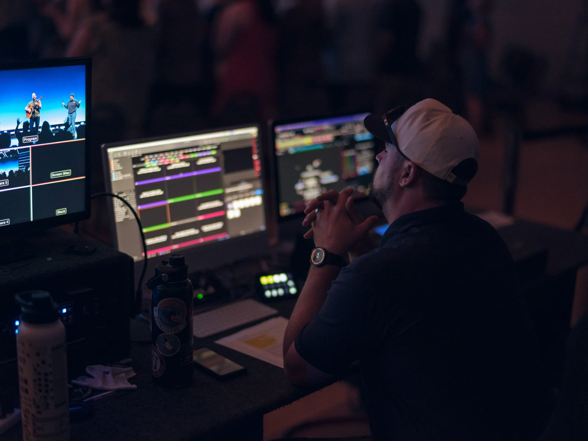 A person wearing a beige baseball cap and glasses is sitting in front of multiple computer screens with colorful editing or production software. There are monitors displaying a stage with two performers, musical instruments, and audience silhouettes. The environment appears dark, indicating a live production, event, or control room setting.