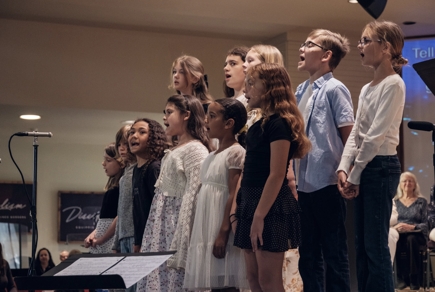 Children singing on stage at a choir performance, holding hands, with audience members in the background.